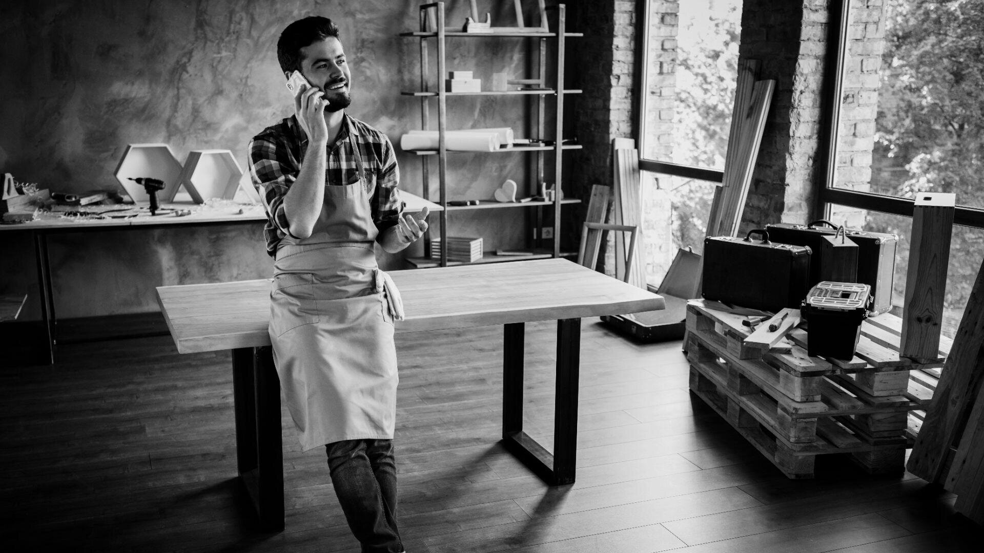 Ein Mann mit einer Schürze steht in einer Werkstatt mit Holztischen, Werkzeugen und Regalen in der Nähe großer Fenster, die natürliches Licht hereinlassen, und telefoniert.