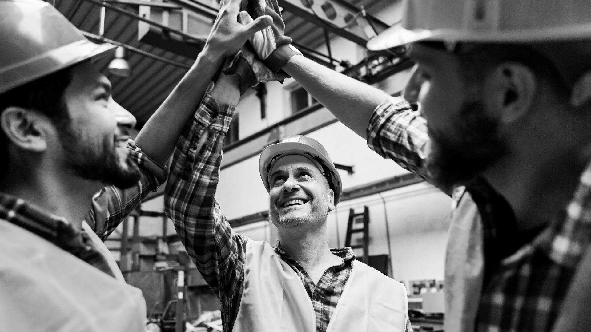 Drei Bauarbeiter mit Schutzhelmen und Sicherheitswesten lächeln und geben sich in einer Werkstatt oder einem Lagerhaus ein High Five und feiern Teamwork und Erfolg.