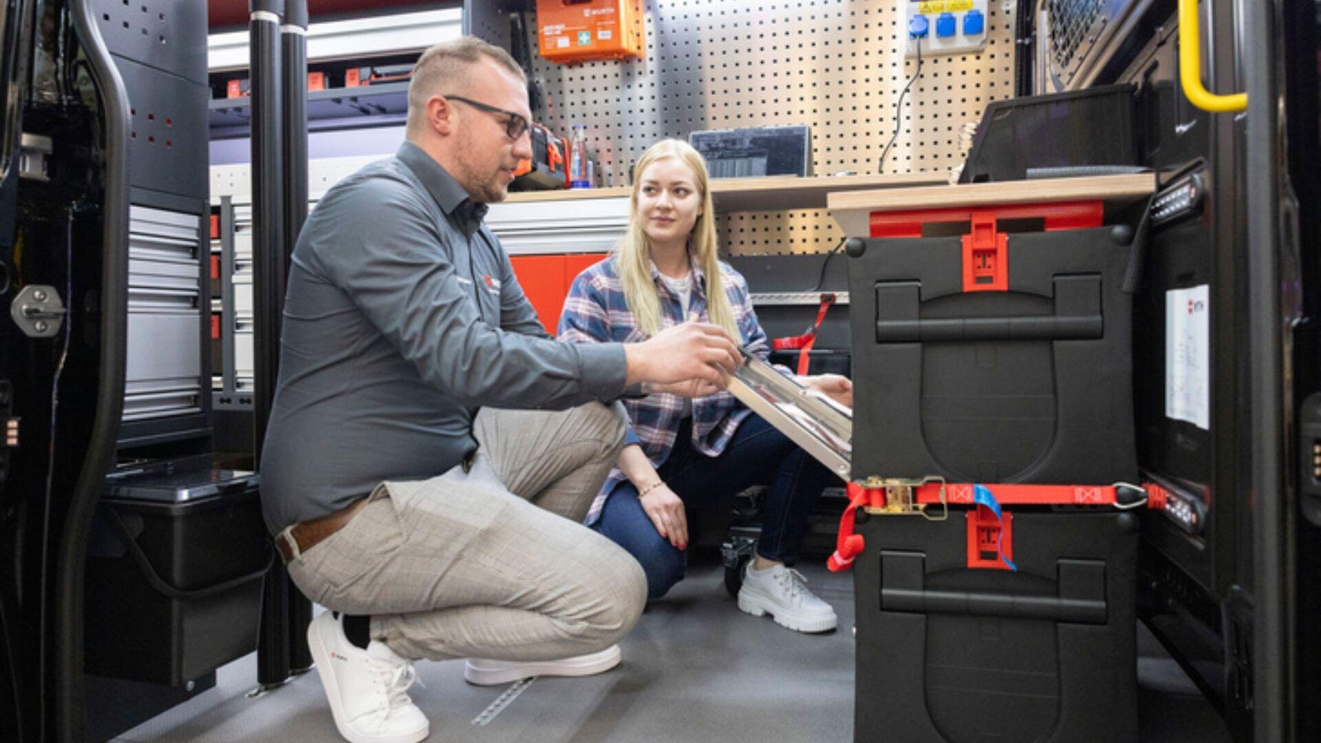 Ein Mann und eine Frau knien neben geordneten Lagerboxen in einer Werkstatt oder einem Lieferwagen, besprechen Werkzeuge oder Geräte und untersuchen dabei ein Klemmbrett.