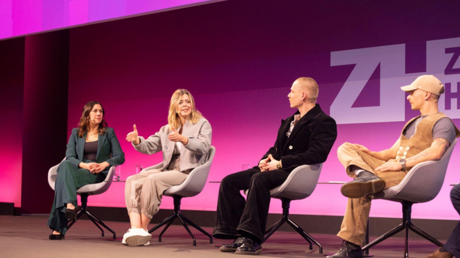 Vier Personen sitzen auf modernen Stühlen auf der Bühne und diskutieren an einer Podiumsdiskussion. Der Hintergrund ist rosa und lila, eine Frau gestikuliert beim Sprechen, während die anderen aufmerksam zuhören.