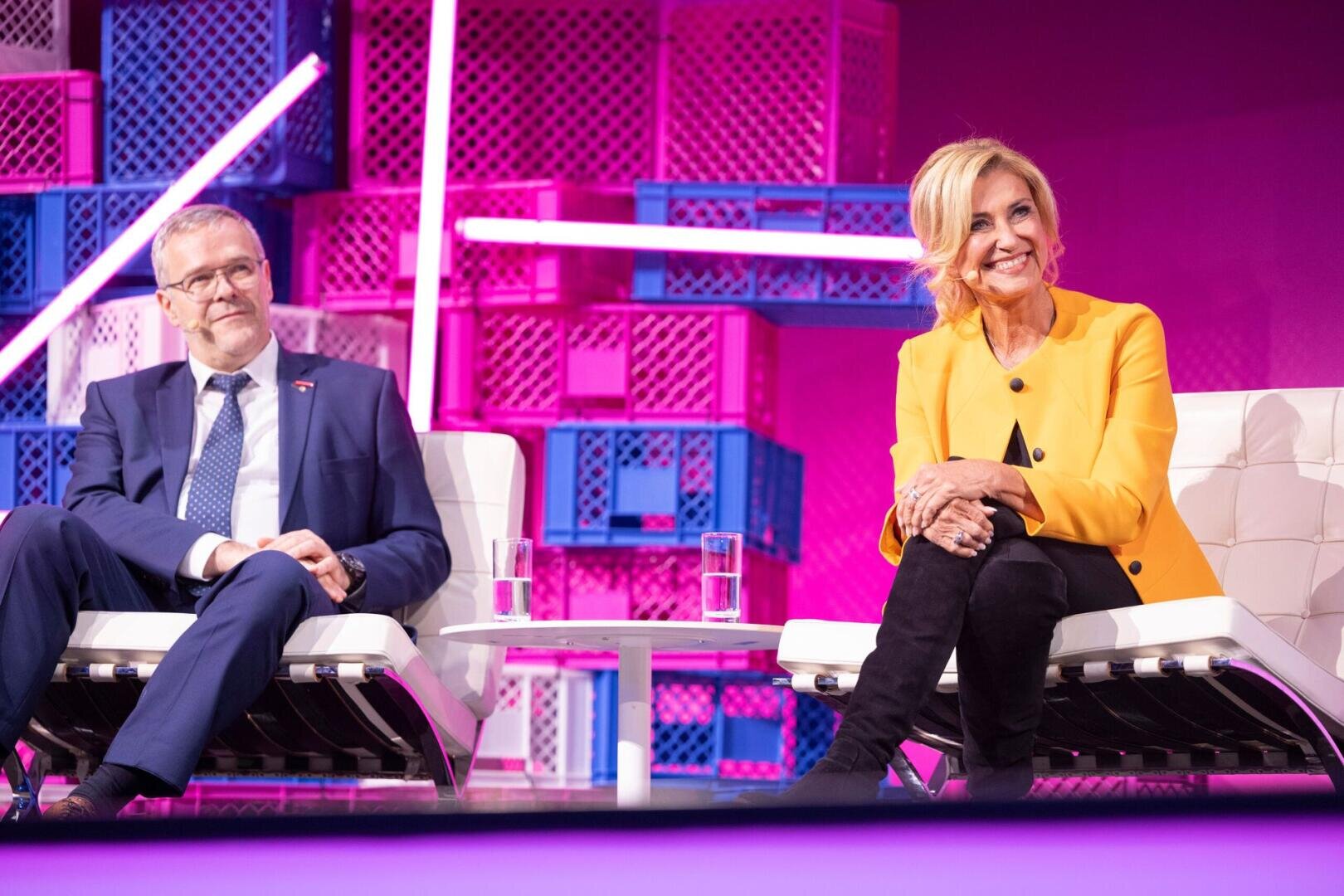 Ein Mann im marineblauen Anzug und eine Frau im leuchtend gelben Blazer sitzen auf weißen Stühlen auf einer hell erleuchteten Bühne mit violettem und rosa Hintergrund, lächeln und hören aufmerksam zu.