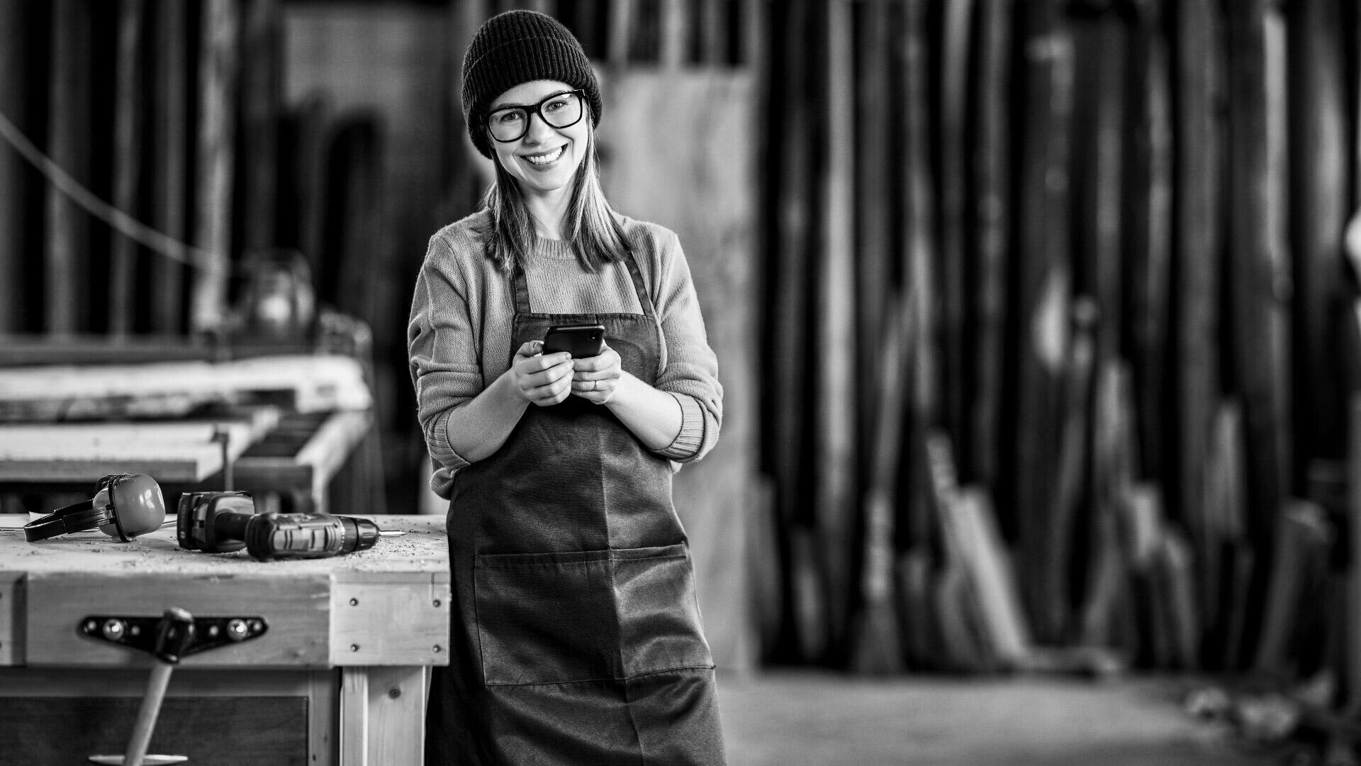Eine Frau, die eine Brille, eine Mütze und eine Schürze trägt, lächelt und hält ein Smartphone in einer Holzwerkstatt. Um sie herum sind Werkzeuge und Holzstücke zu sehen. Das Foto ist in schwarz-weiß gehalten.