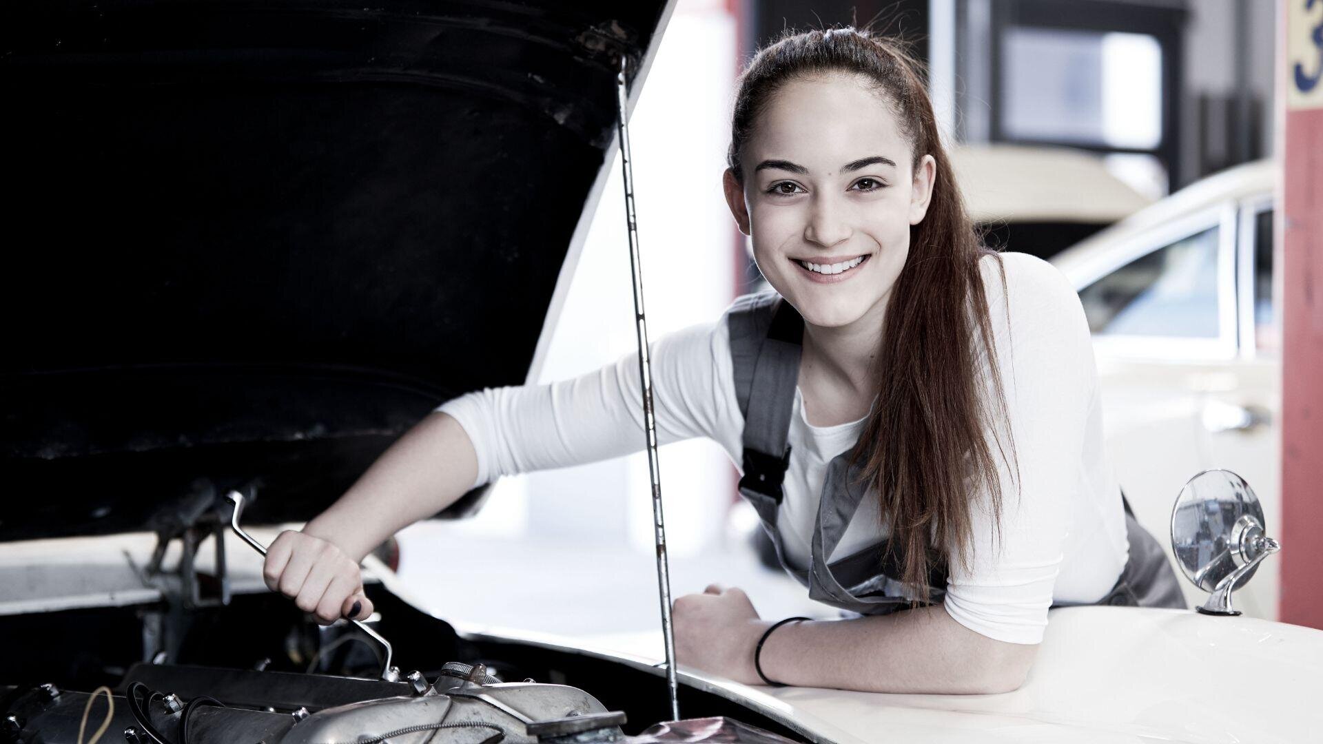 Eine lächelnde junge Frau im Overall arbeitet bei geöffneter Motorhaube an einem Automotor und hält in einer hell erleuchteten Garage ein Werkzeug in der Hand.