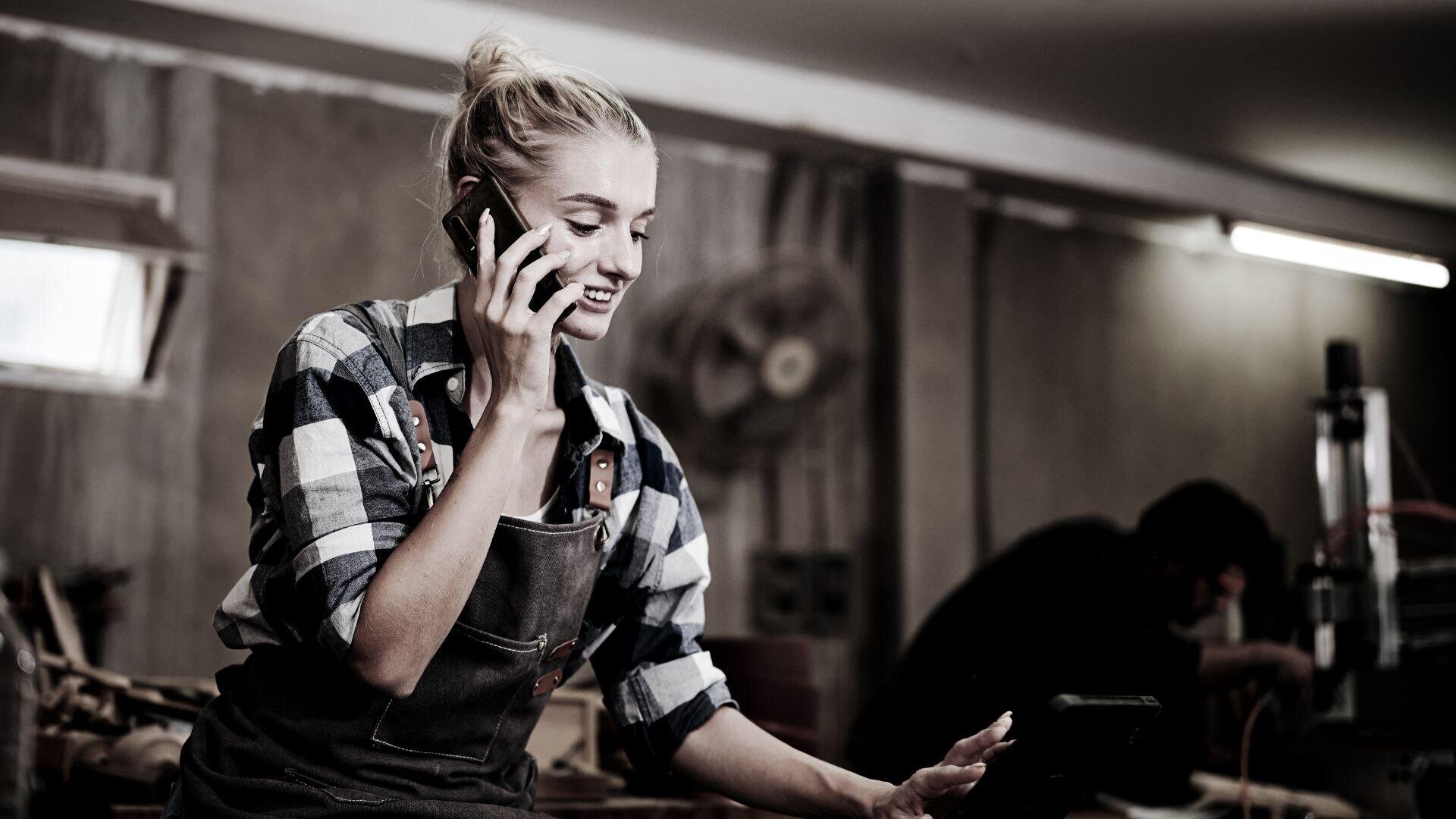 Eine Frau in kariertem Hemd und Schürze lächelt, während sie in einer Werkstatt mit einem Smartphone spricht. Im Hintergrund sind Werkzeuge und eine verschwommene Figur zu sehen.