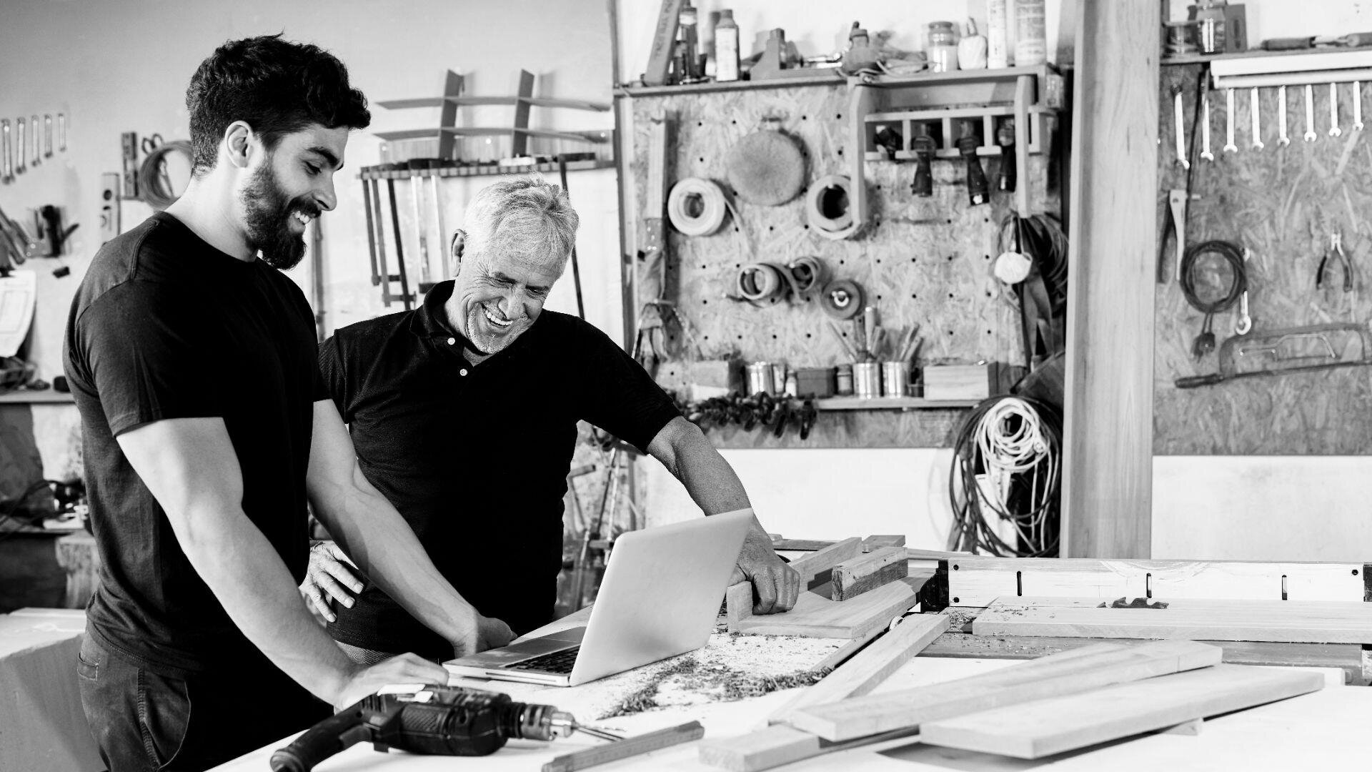 Zwei Männer stehen lächelnd in einer Werkstatt und schauen auf einen Laptop auf einer Werkbank, die mit Holzstücken und Werkzeugen bedeckt ist. Im Hintergrund sind Regale und Hängevorrichtungen zu sehen.