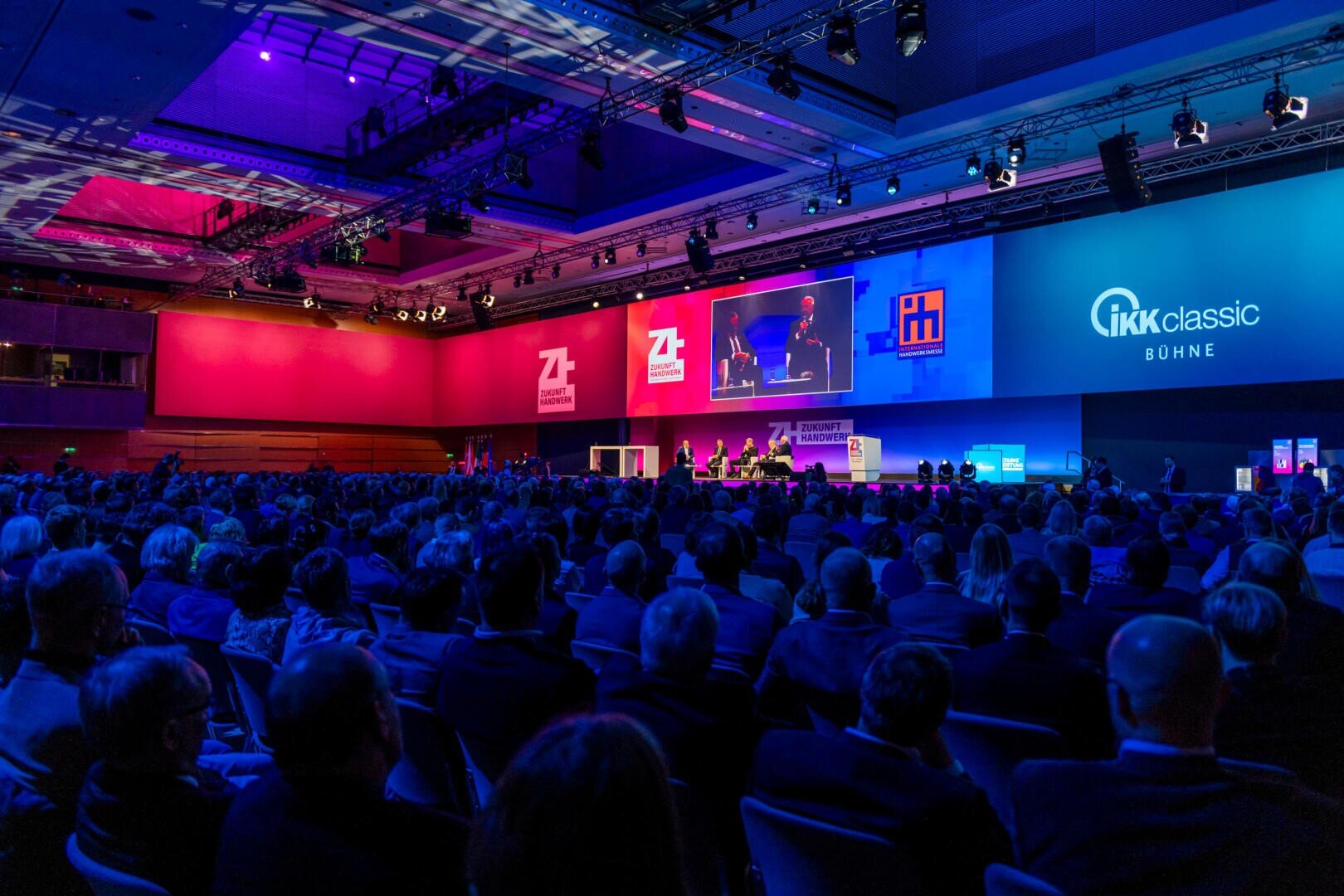 Ein großes Publikum nimmt an einer Konferenz in einem geräumigen, modernen Auditorium mit bunter Beleuchtung, einer Podiumsdiskussion und projizierten Logos an den Wänden teil.