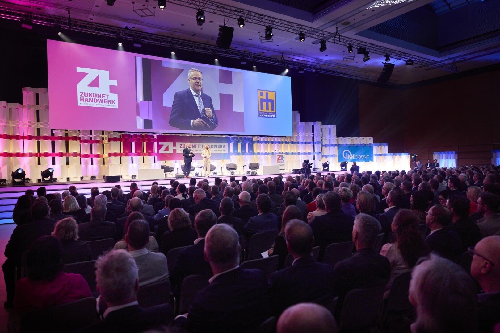 Ein Redner steht auf der Bühne und spricht zu einem großen, sitzenden Publikum auf einer Konferenz. Hinter ihm befinden sich helle Lichter, bunte Banner und ein großer Bildschirm, auf dem der Redner und die Veranstaltungslogos angezeigt werden.