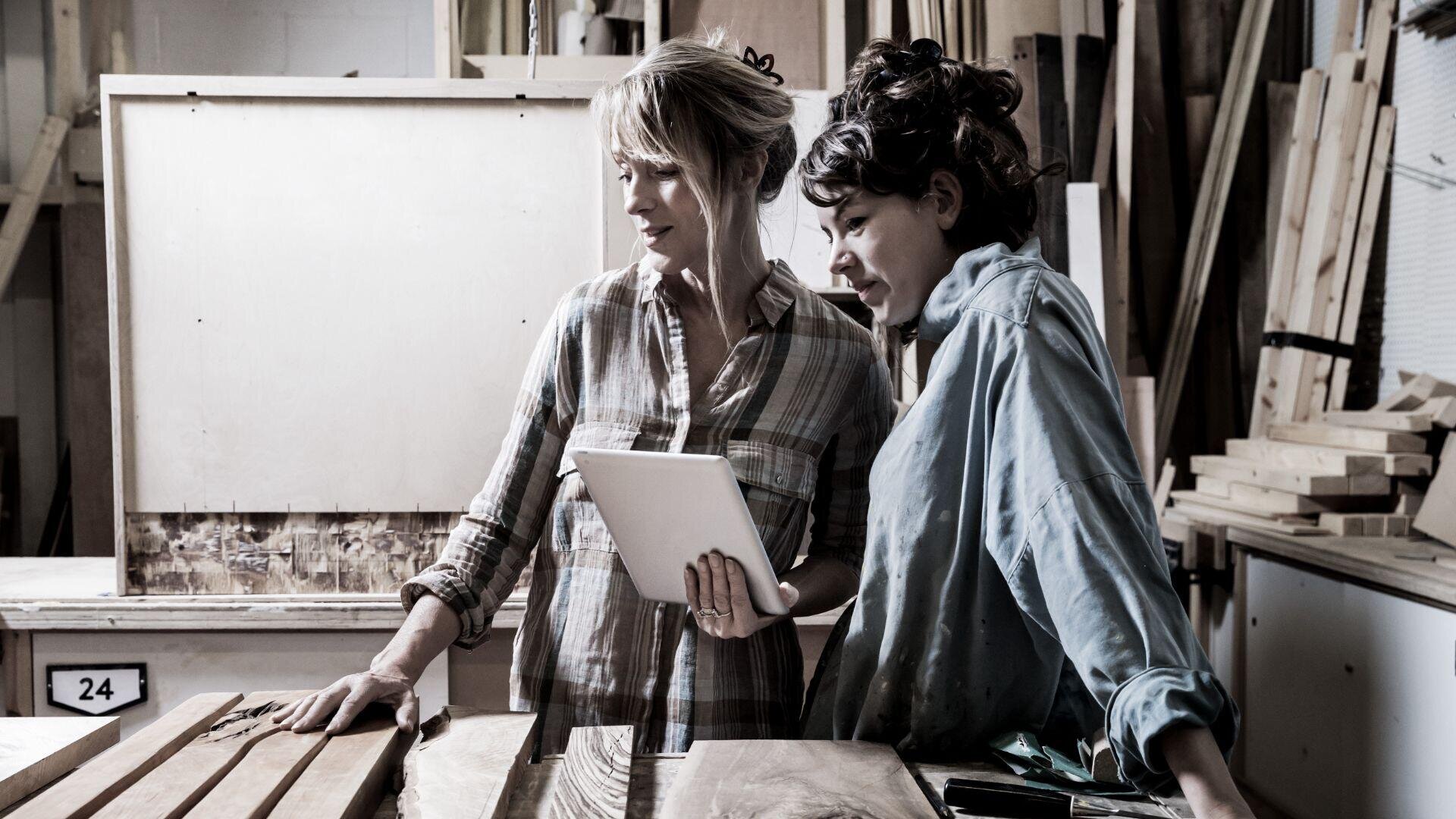 Zwei Frauen in einer Holzwerkstatt untersuchen ein Stück Holz auf einem Tisch. Eine hält ein Tablet in der Hand, beide tragen legere Arbeitskleidung, umgeben von verschiedenen Holzbrettern und Werkstattwerkzeugen.