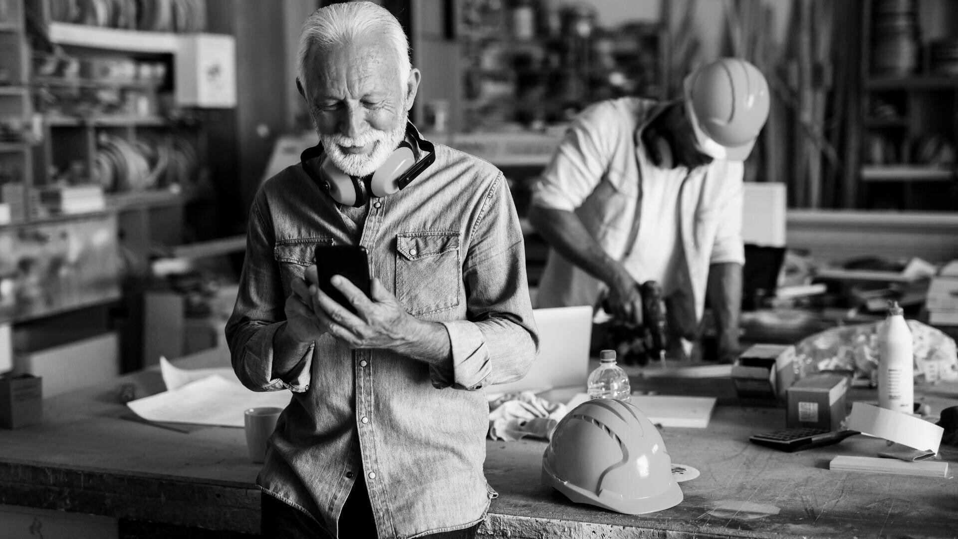 Ein älterer Mann mit grauem Haar und Kopfhörern um den Hals schaut auf sein Telefon, während er an einer Werkbank steht; eine andere Person mit Schutzhelm arbeitet im Hintergrund, umgeben von Werkzeugen und Papieren.