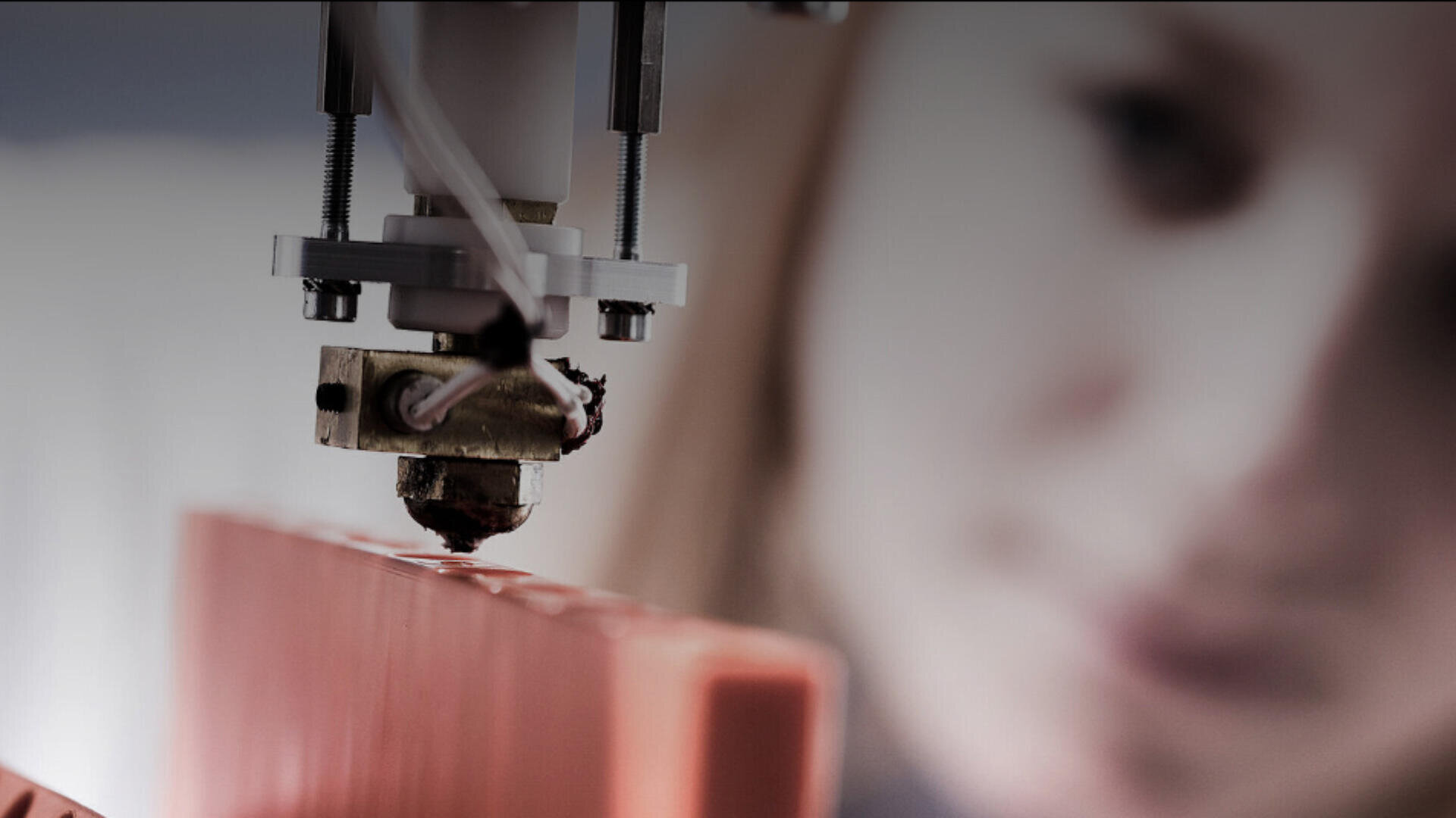 Nahaufnahme einer 3D-Druckerdüse, die ein orangefarbenes Objekt druckt, mit einer verschwommenen Person im Hintergrund, die den Vorgang aufmerksam beobachtet.