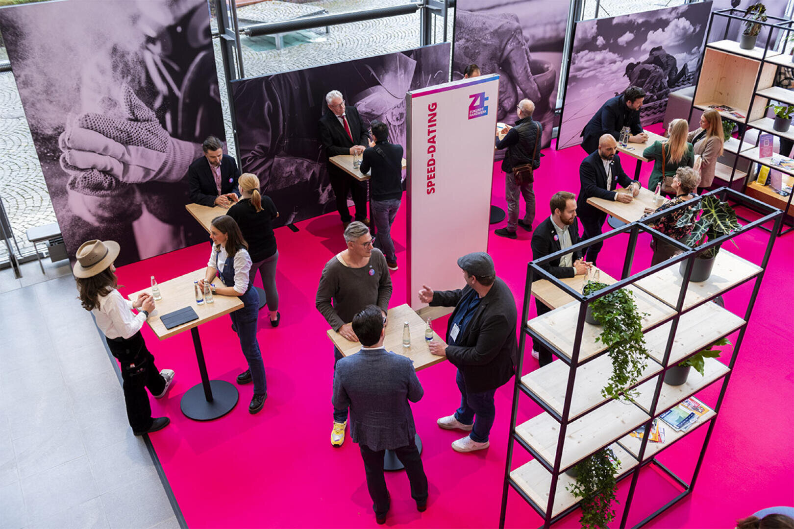 Während einer Speed-Dating-Veranstaltung in einem hellen, modernen Raum mit rosafarbenem Boden unterhalten sich Menschen an hohen Tischen. Große Schwarz-Weiß-Fotos und Regale mit Pflanzen schmücken den Raum.