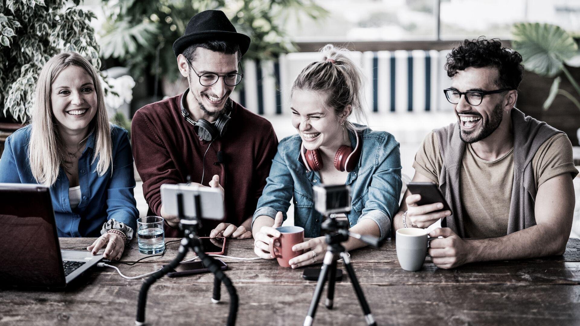Vier junge Erwachsene sitzen mit Laptops, Kaffeetassen und einem Smartphone an einem Holztisch und nehmen lachend gemeinsam ein Video auf. Vor ihnen sind zwei Kameras auf kleinen Stativen aufgebaut.