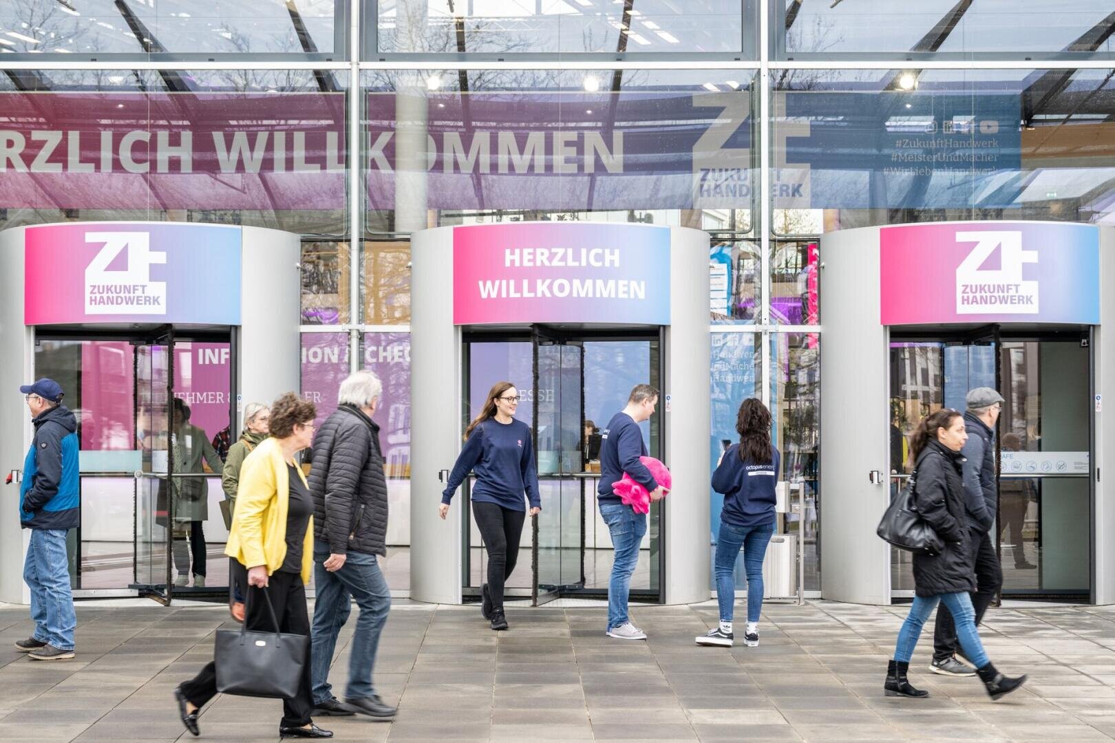 Menschen gehen vorbei und betreten durch eine Glasdrehtür unter einem Schild mit der Aufschrift „Herzlich Willkommen“ den Eingang eines modernen Gebäudes zur Veranstaltung „Zukunft Handwerk“. Die Szene ist hell und belebt, viele Passanten strömen vorbei.