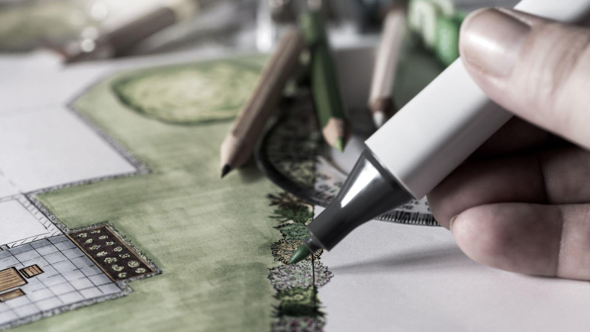 Eine Hand verwendet einen grünen Marker, um eine Landschaftsentwurfszeichnung auszumalen, während Buntstifte und weitere Zeichenwerkzeuge auf dem Papier liegen.
