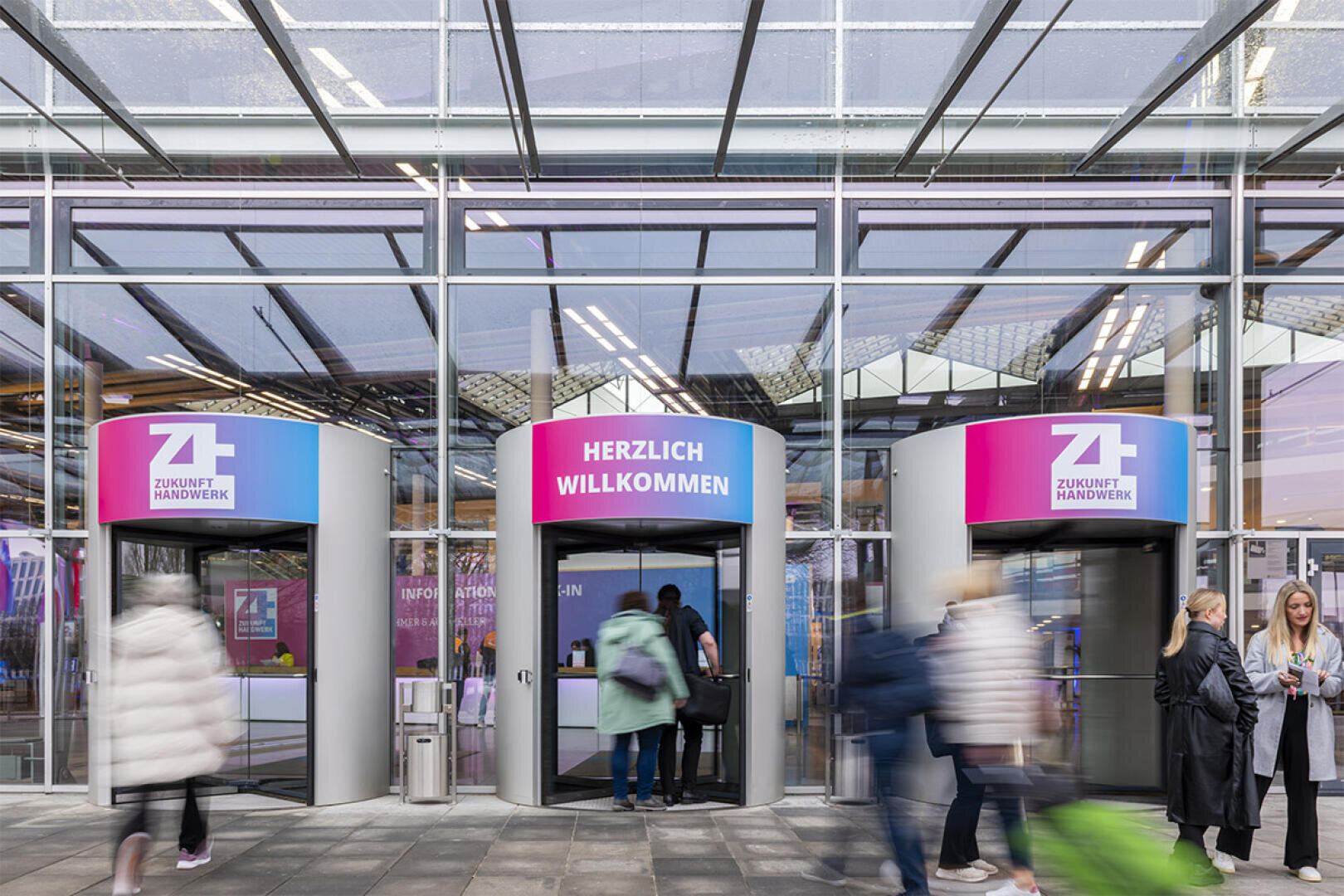 In einem modernen Glasgebäude gehen Menschen durch Drehtüren ein und aus. Über den Türen sind bunte Schilder angebracht, eines mit der Aufschrift HERZLICH WILLKOMMEN (Deutsch für Willkommen). Mehrere Menschen gehen oder stehen in der Nähe.