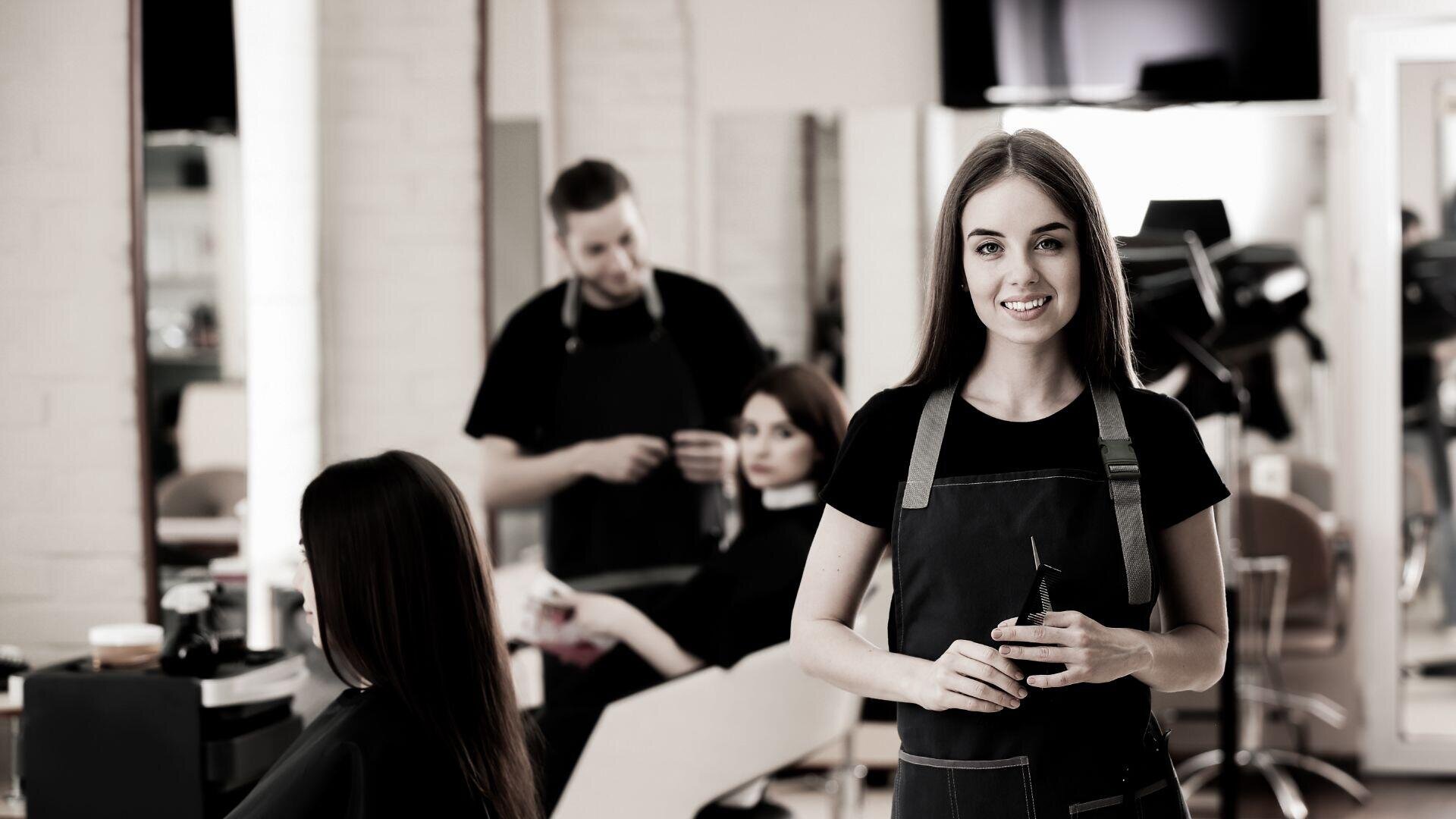 Eine Frau in einer schwarzen Schürze steht lächelnd mit einer Schere in der Hand in einem Friseursalon. Hinter ihr bearbeitet ein Friseur die Haare einer sitzenden Kundin. Im Hintergrund sind Spiegel und Friseurstühle zu sehen.