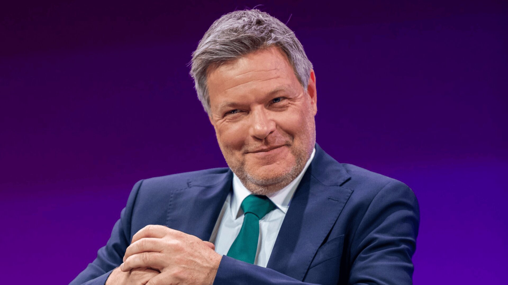 Ein Mann in marineblauem Anzug, weißem Hemd und grüner Krawatte sitzt auf einem weißen Stuhl vor violettem Hintergrund. Er lächelt leicht und hat die Hände gefaltet. Vor ihm steht ein runder weißer Tisch.