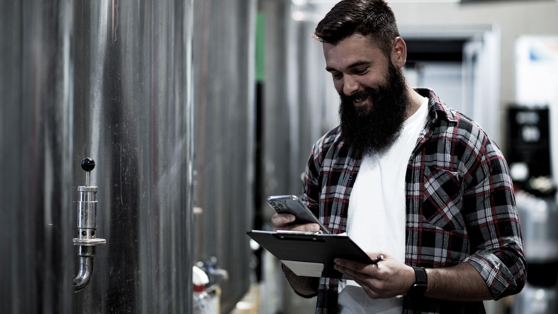 Ein bärtiger Mann in einem karierten Hemd steht lächelnd in einer Brauerei, während er auf sein Telefon schaut und ein Klemmbrett in der Hand hält. Im Hintergrund sind große Edelstahltanks zu sehen.