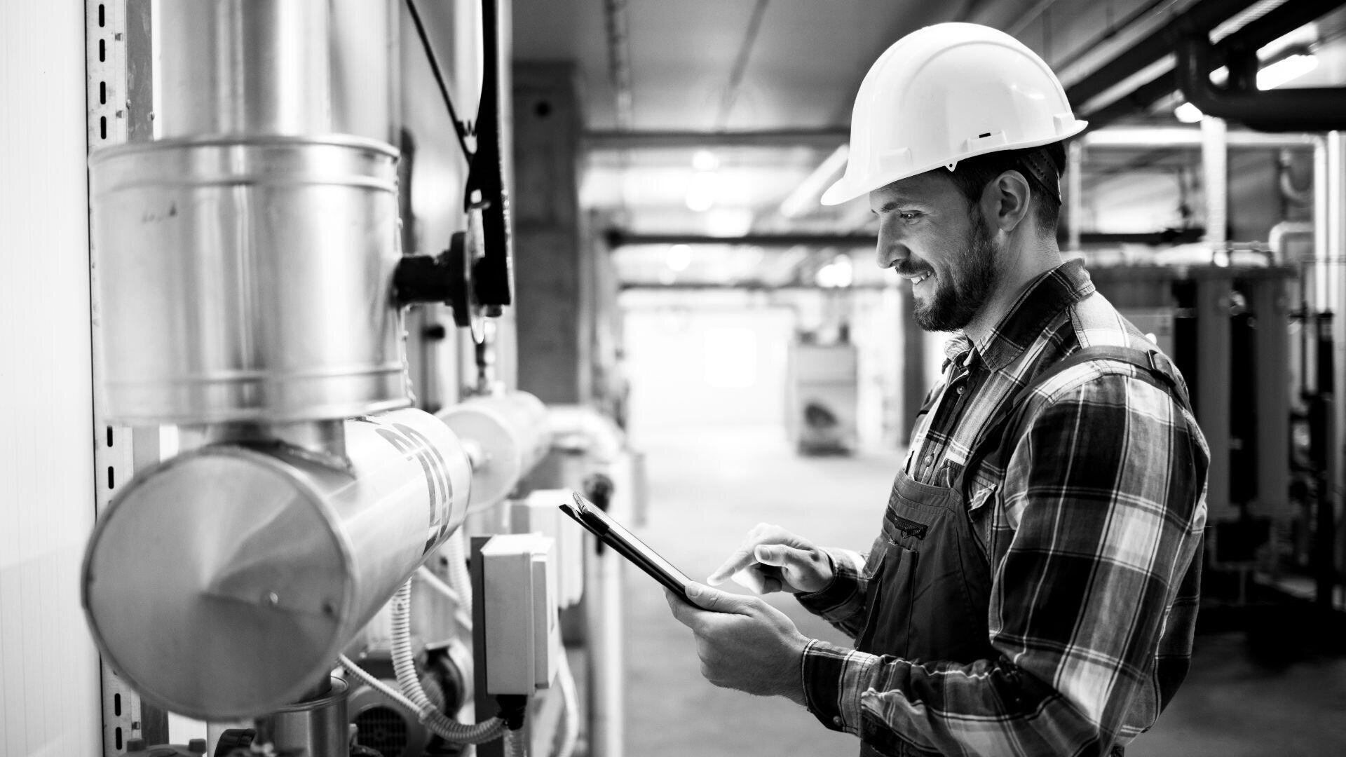 Ein Mann mit Schutzhelm und kariertem Hemd arbeitet in einer Industrieanlage, lächelt, während er Geräte inspiziert und ein Tablet verwendet.