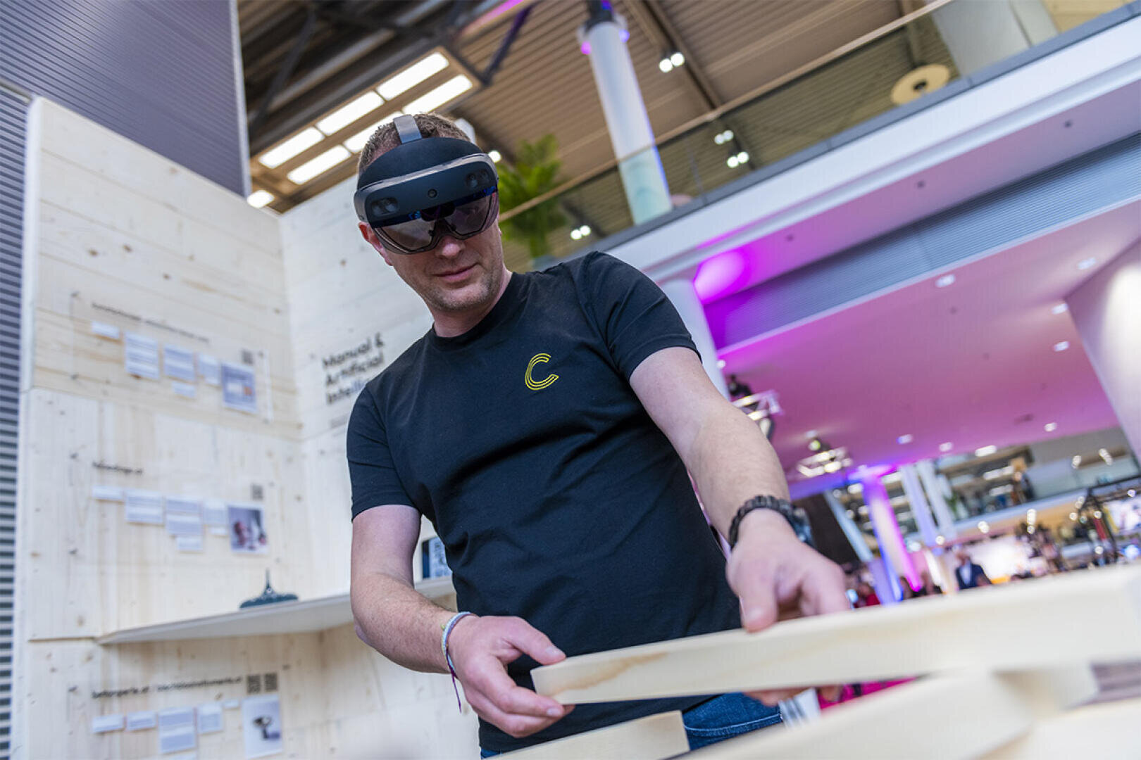 Ein Mann mit einer Augmented-Reality-Brille interagiert mit Holzbrettern in einem modernen, hell beleuchteten Innenraum mit hoher Decke und zeitgenössischem Dekor.