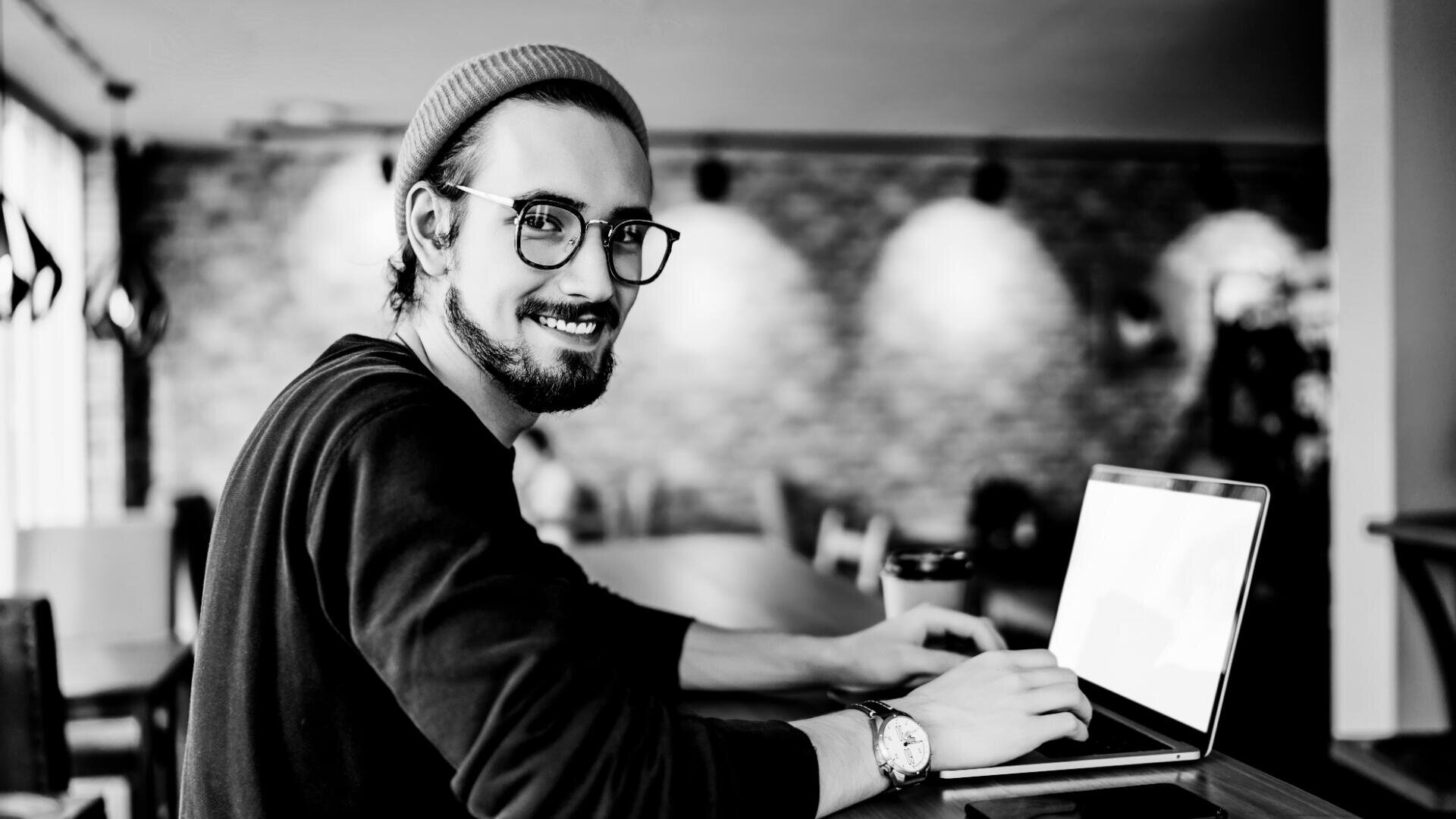 Eine lächelnde Person mit Brille und Beanie sitzt an einem Tisch und tippt auf einem Laptop. Neben ihr steht eine Kaffeetasse. Im Hintergrund ist ein verschwommenes Café mit Backsteinmauern zu sehen.
