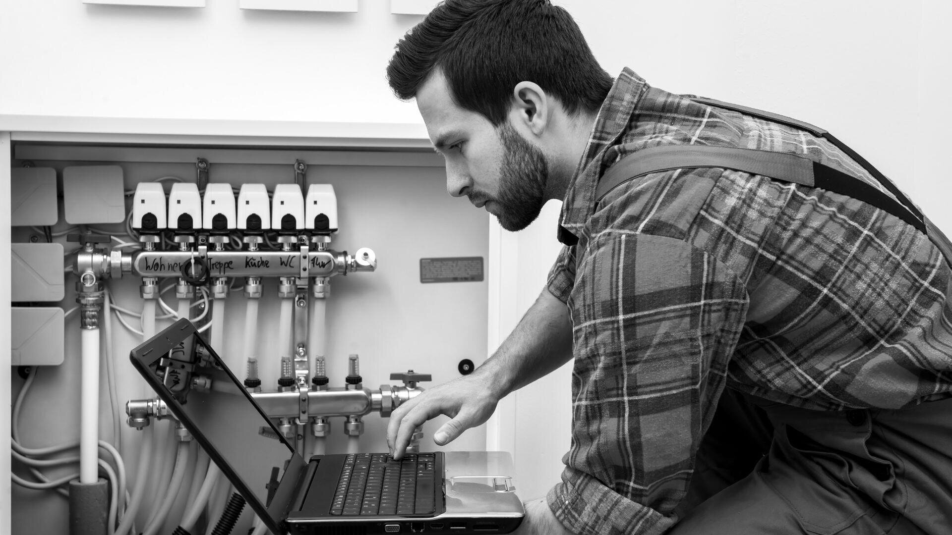 Ein Mann in kariertem Hemd und Overall kniet neben offenen Sanitäranlagen und führt mit einem Laptop Diagnosearbeiten durch. Es scheint sich um eine Wartungs- oder technische Inspektionsszene in einem Innenbereich zu handeln.