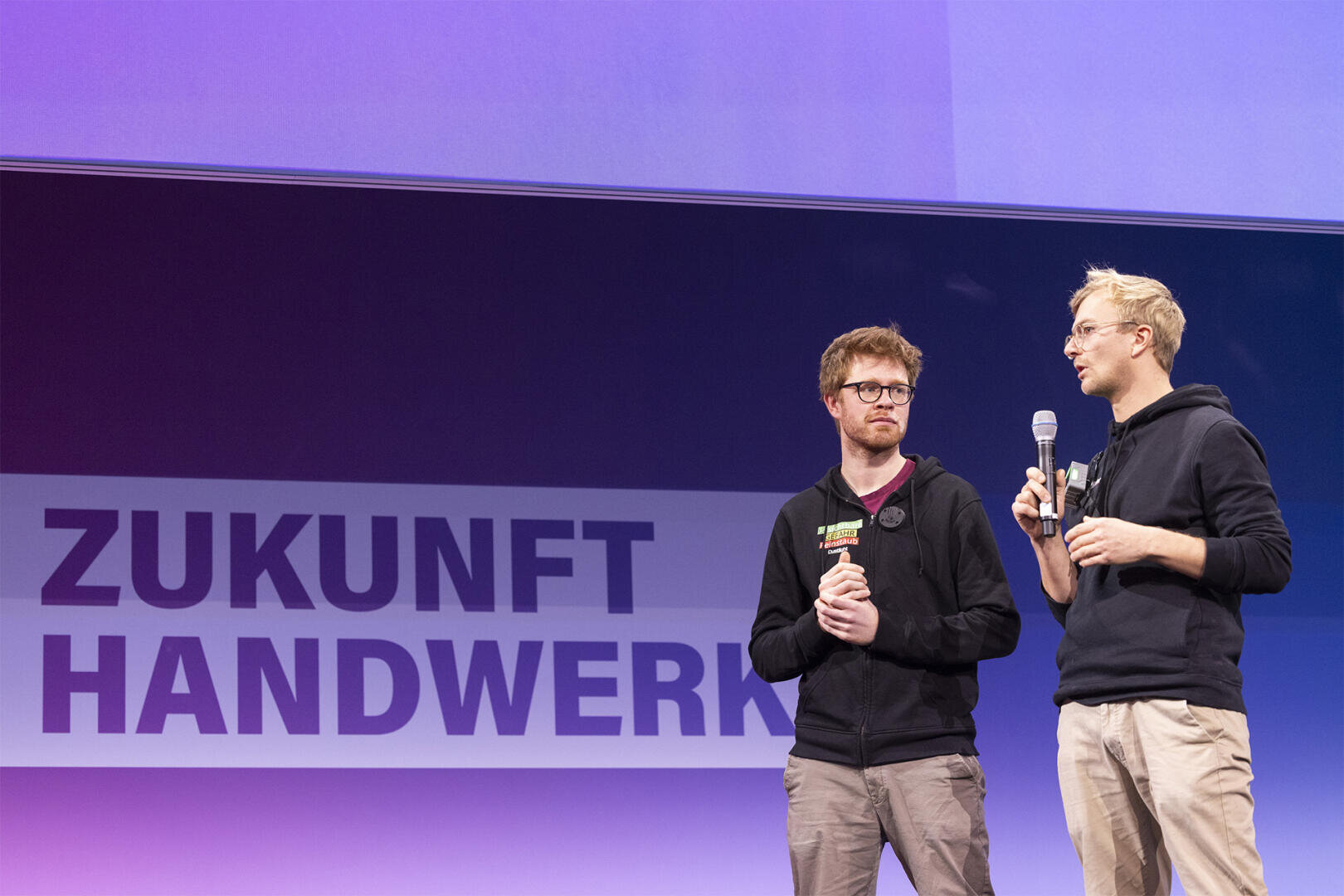 Zwei Männer in Freizeitkleidung stehen auf der Bühne mit Mikrofonen vor einem großen Schild mit der Aufschrift ZUKUNFT HANDWERK vor einem lila beleuchteten Hintergrund.