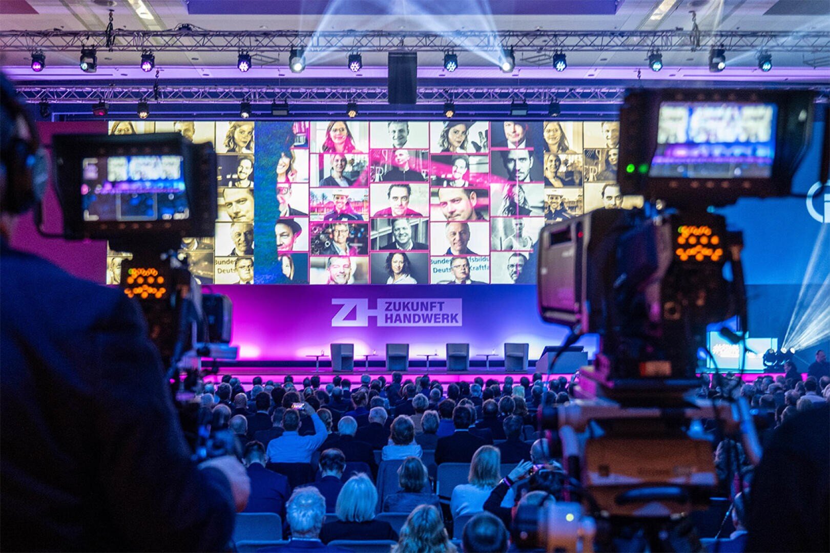Ein großes Publikum blickt auf eine Bühne mit vier leeren Stühlen und einer großen Leinwand, auf der viele Gesichter bei einer Veranstaltung von Zukunft Handwerk zu sehen sind.