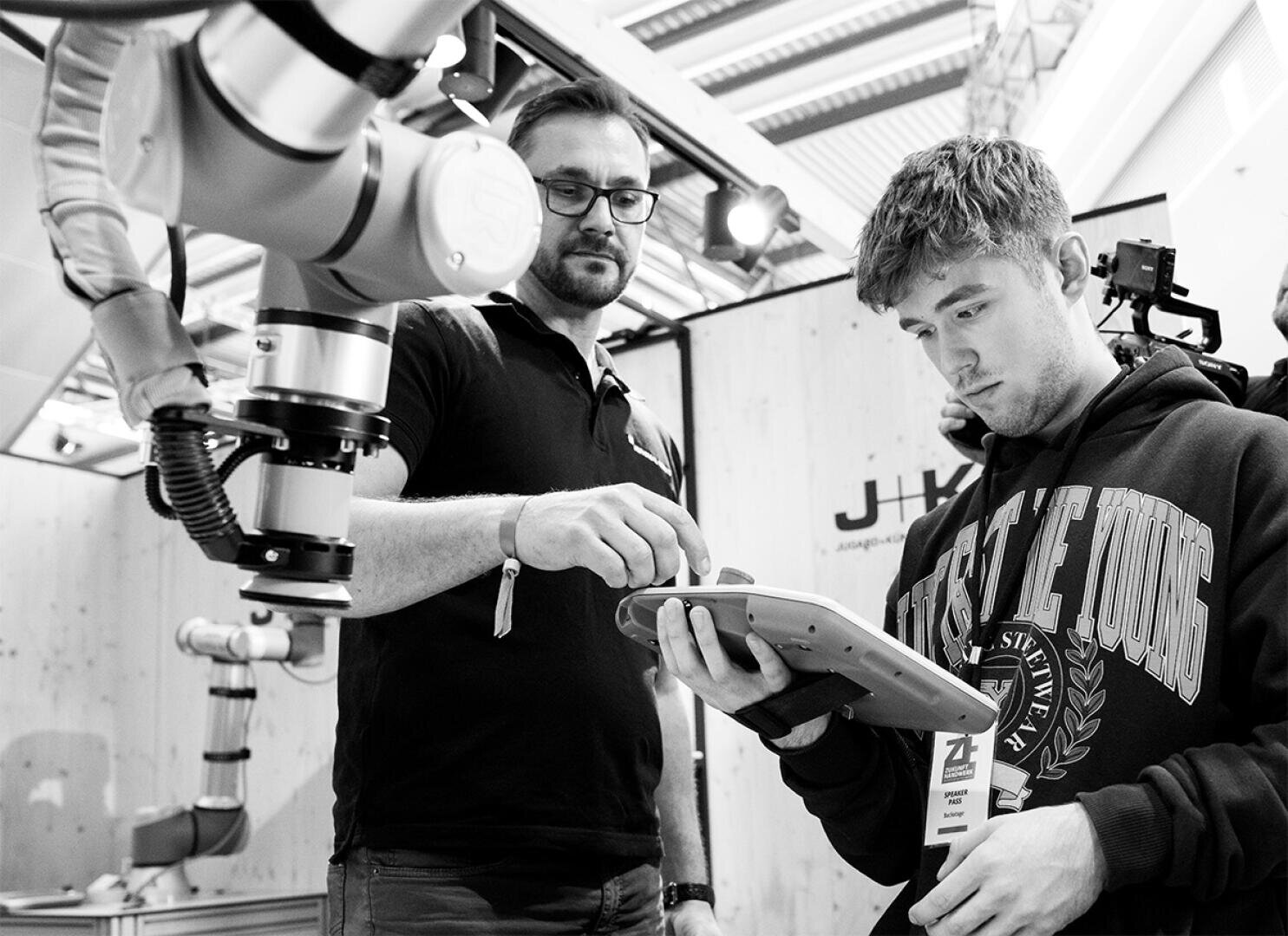 Zwei Männer interagieren mit einem Roboterarm; einer zeigt, während der andere ein Touchscreen-Tablet verwendet, was auf eine Demonstration oder Zusammenarbeit in einem modernen, technikorientierten Umfeld hindeutet.