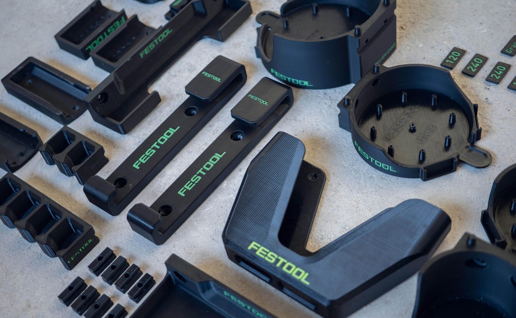 Auf einer hellen Fläche sind mehrere schwarze Festool Werkzeugzubehörteile aus Kunststoff mit grünen Etiketten übersichtlich angeordnet und zeigen Teile in verschiedenen Formen und Größen.