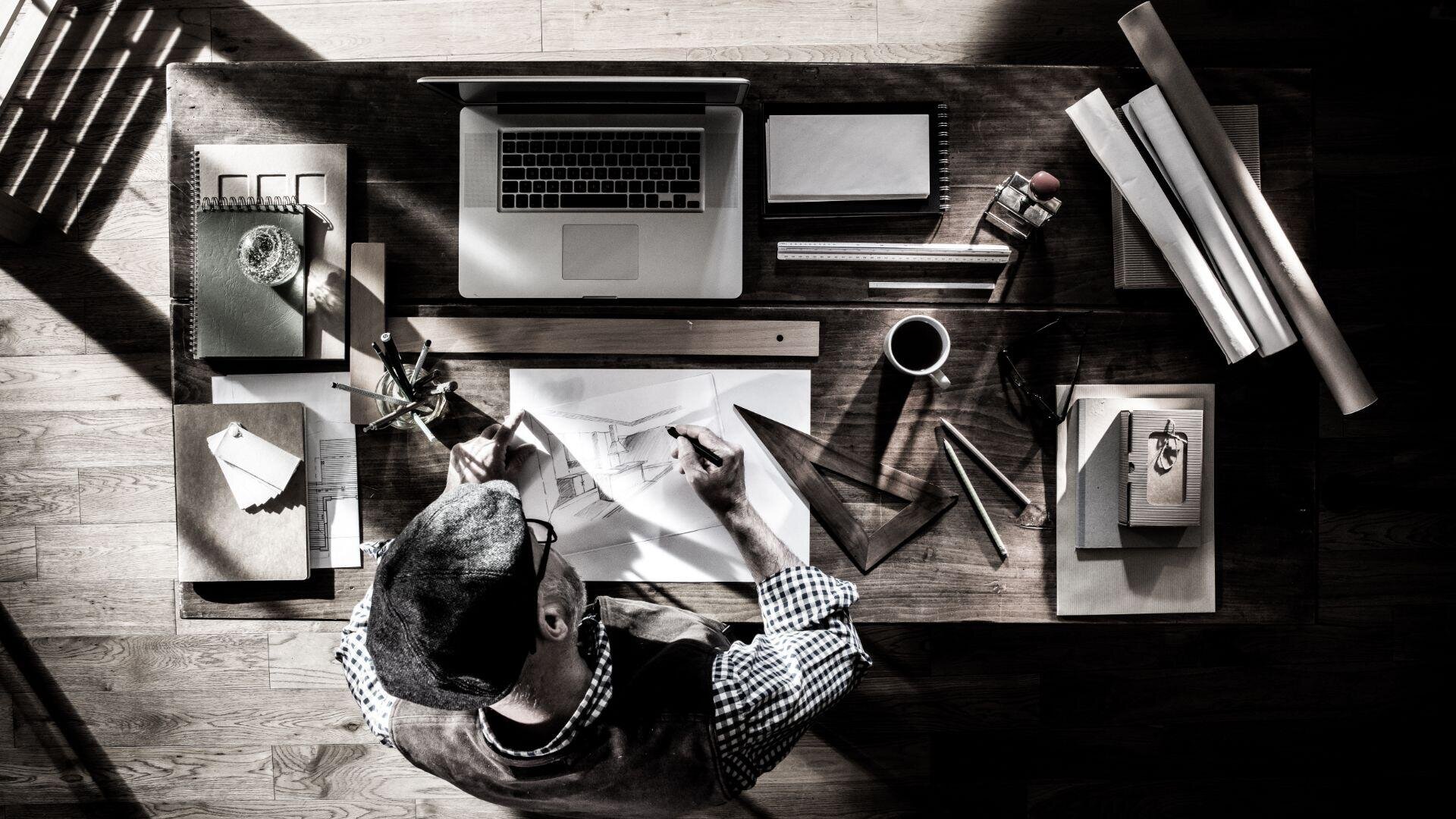 Eine Draufsicht auf eine Person, die an einem Holzschreibtisch mit Laptop, Kaffeetasse, Notizbüchern, Linealen, Bleistiften und zusammengerollten Papieren Architekturpläne skizziert, beleuchtet vom Sonnenlicht, das durch ein Fenster fällt.