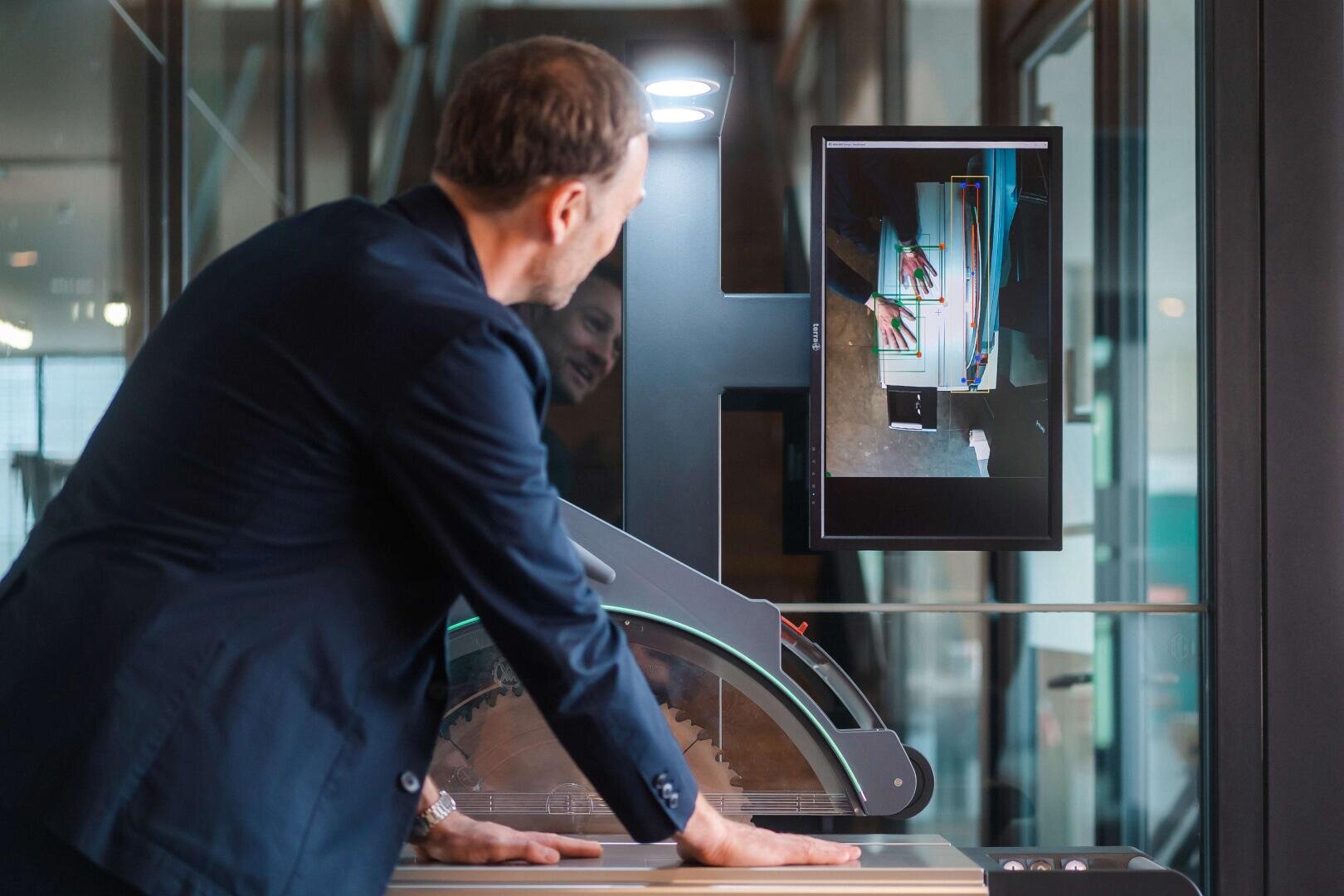 Ein Mann im Anzug untersucht ein digitales Display, das einen detaillierten Scan oder ein Röntgenbild eines Gemäldes zeigt. Dazu wird ein spezielles Scangerät in einem modernen, verglasten Raum verwendet.