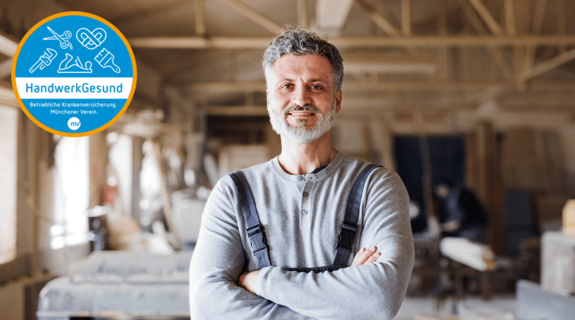 Ein Mann mittleren Alters mit grauem Haar und Bart, der eine Latzhose und ein graues Hemd trägt, steht lächelnd mit verschränkten Armen in einer Holzwerkstatt. In der oberen linken Ecke ist ein blaues HandwerkGesund-Abzeichen zu sehen.