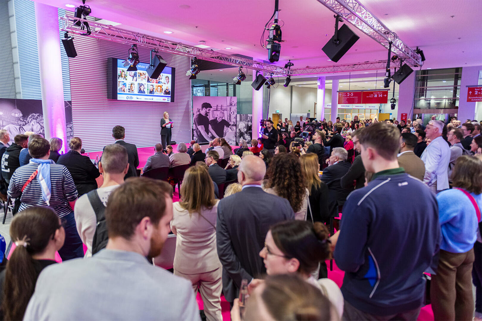 Eine große Gruppe von Menschen verfolgt eine Präsentation in einem hell erleuchteten Raum mit rosa Licht. Ein Redner steht auf der Bühne neben einem Bildschirm, der Bilder zeigt. Das Publikum sitzt und steht mit Blick auf die Bühne.