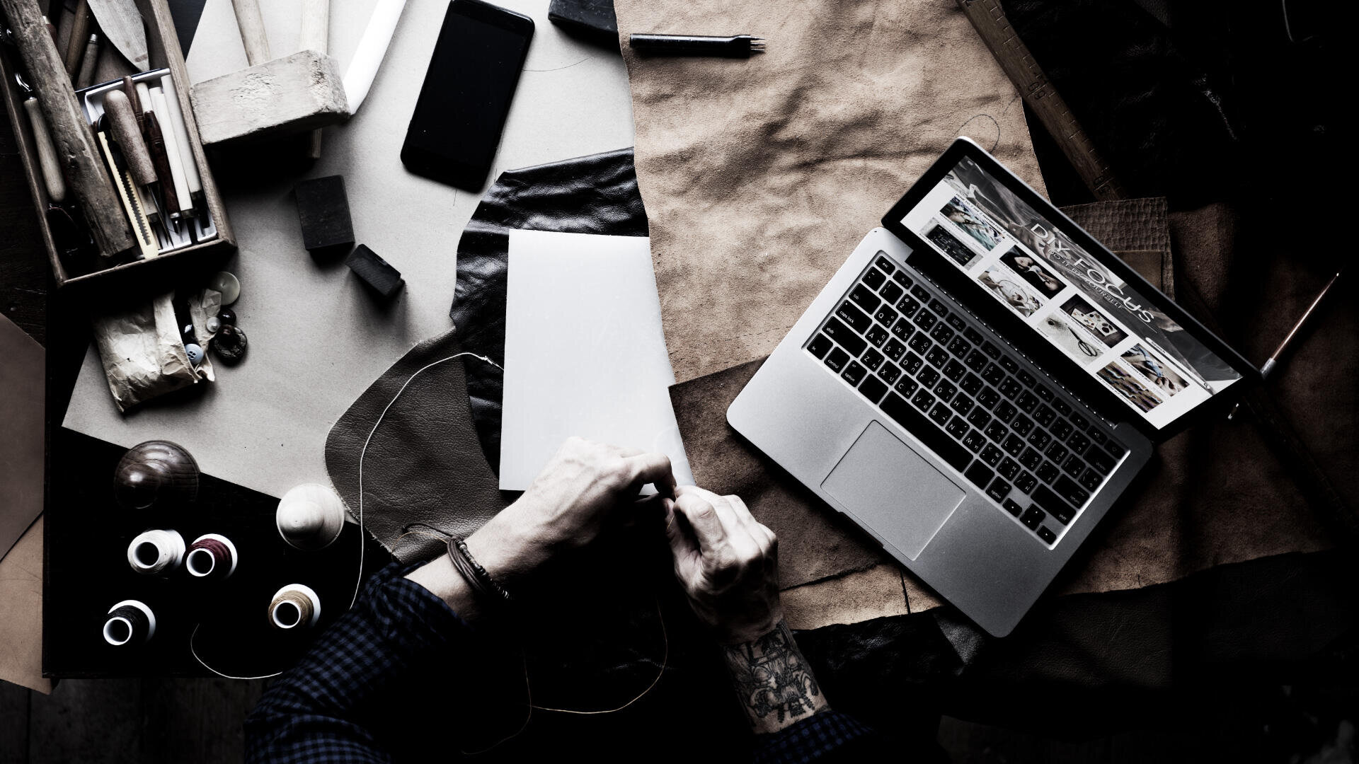 Eine Person arbeitet mit Lederstücken an einem überfüllten Schreibtisch, umgeben von Werkzeugen, Garnrollen, einem Smartphone und einem offenen Laptop, auf dem eine Website angezeigt wird.