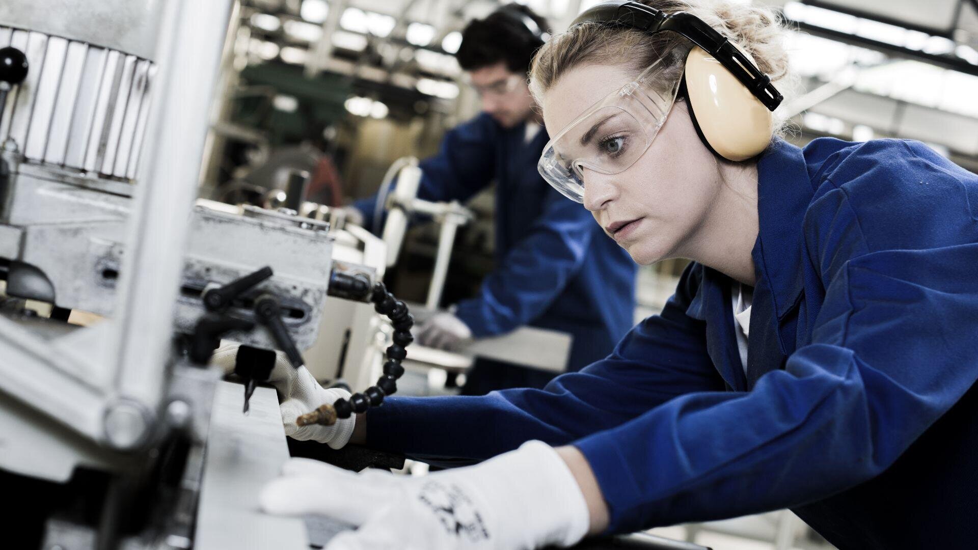 Eine Frau mit Schutzbrille, Ohrenschützern, Handschuhen und blauer Uniform bedient Maschinen in einer Fabrik, während im Hintergrund ein weiterer Arbeiter zu sehen ist.