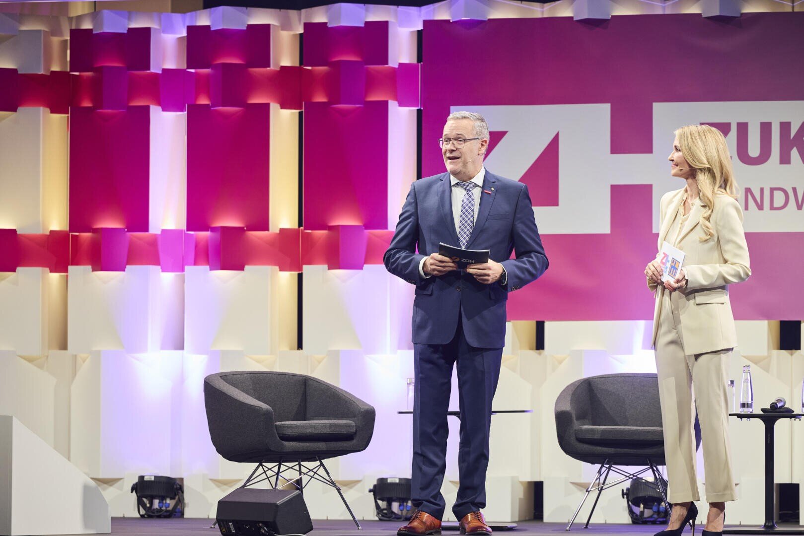 Zwei professionell gekleidete Moderatoren, ein Mann im Anzug und eine Frau in einem hellen Anzug, stehen auf einer Bühne mit modernen Stühlen und einem farbenfrohen geometrischen Hintergrund und sprechen auf einer Veranstaltung oder Konferenz.