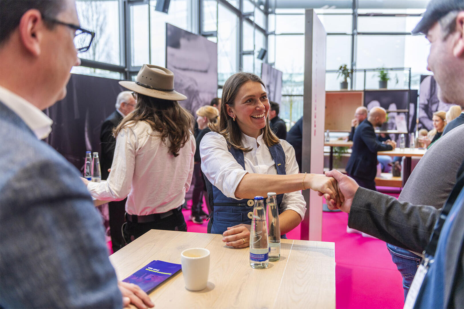 Eine Frau lächelt und schüttelt einem Mann die Hand, der an einem Holztisch sitzt, während sich im Hintergrund mehrere Personen unter die Gäste mischen. Auf dem Tisch stehen eine Kaffeetasse, eine Glaswasserflasche und ein Smartphone.