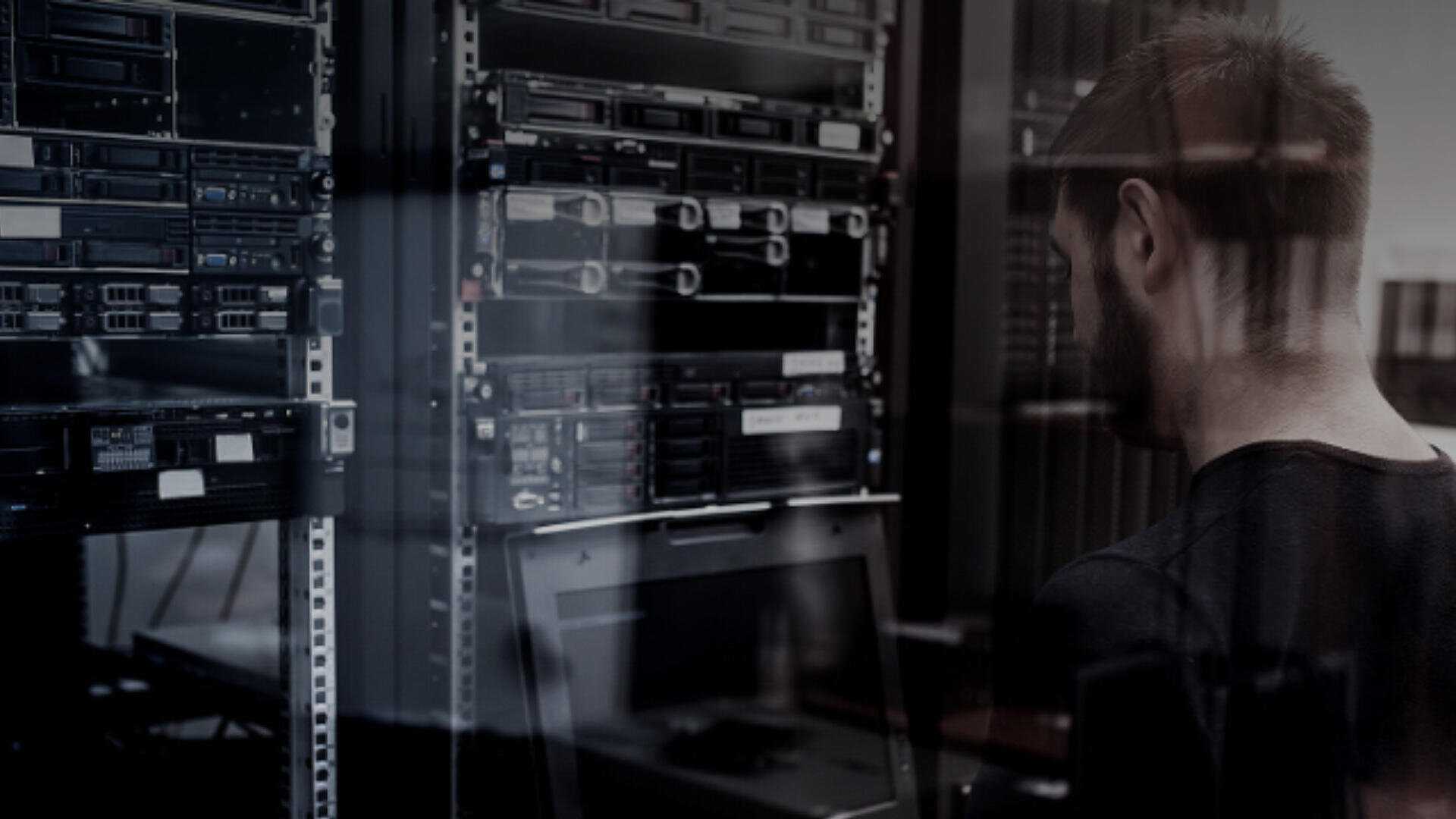 Eine Person steht in einem schwach beleuchteten Raum vor Server-Racks und arbeitet an einem Laptop oder einer Konsole. Auf dem Glas sind Reflexionen sichtbar.