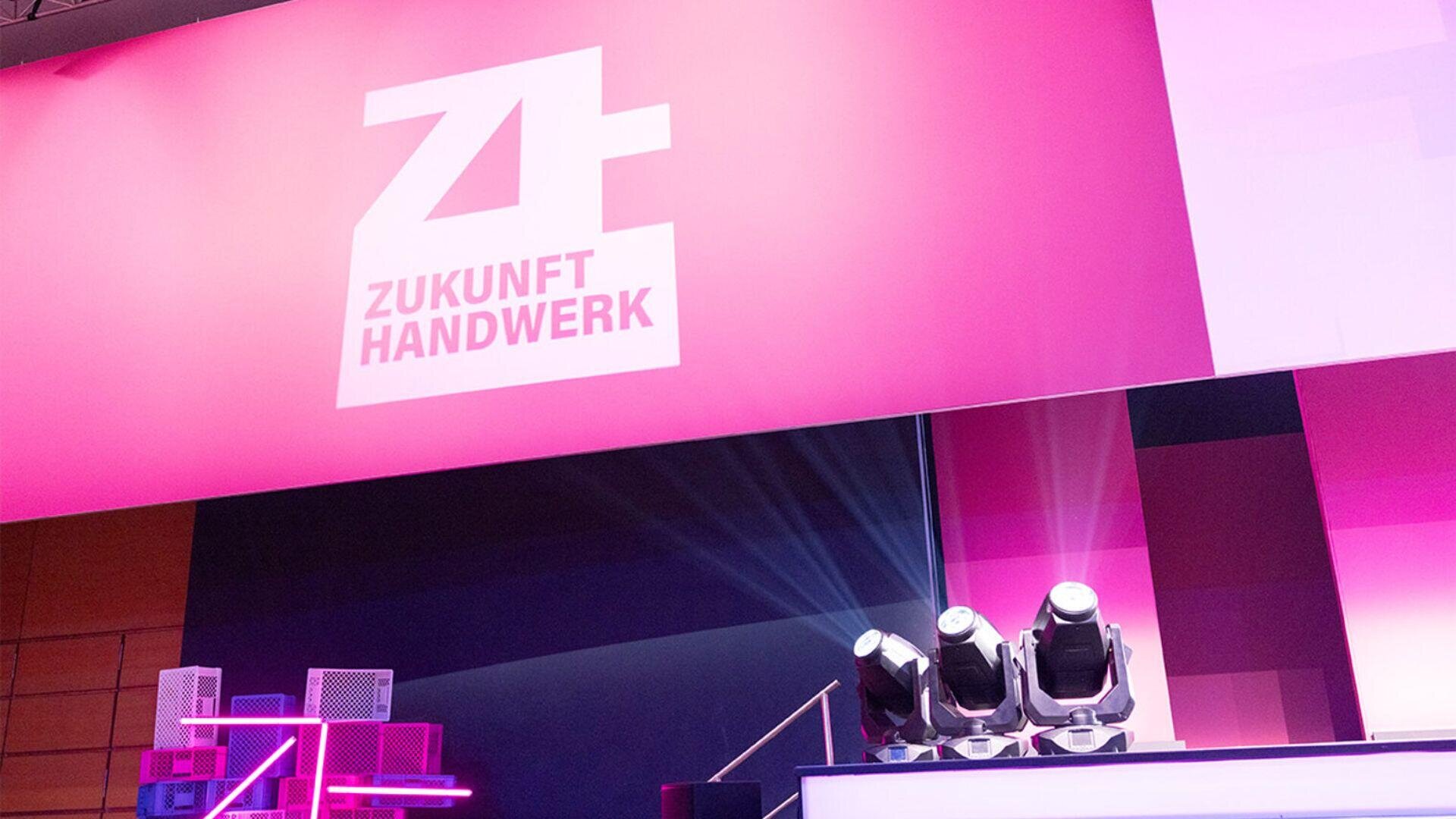 Eine hell erleuchtete Bühne mit einem großen rosa Banner, auf dem das weiße Logo und der Text „Zukunft Handwerk“ zu sehen sind. Die Bühnenlichter sind nach oben gerichtet, im Hintergrund ist modernes Dekor zu sehen.