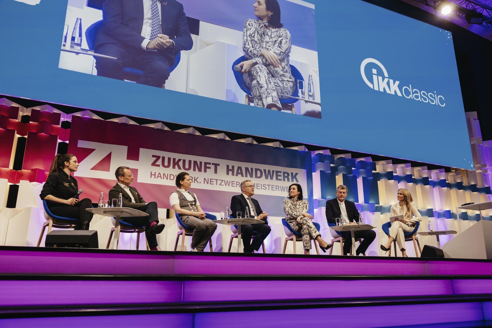 Sechs Personen sitzen auf einer hell erleuchteten Bühne bei einer Podiumsdiskussion im Rahmen der Veranstaltung Zukunft Handwerk. Hinter ihnen ein klassisches IKK-Logo und die Veranstaltungsbeschilderung. Die Kulisse ist modern und bunt beleuchtet.