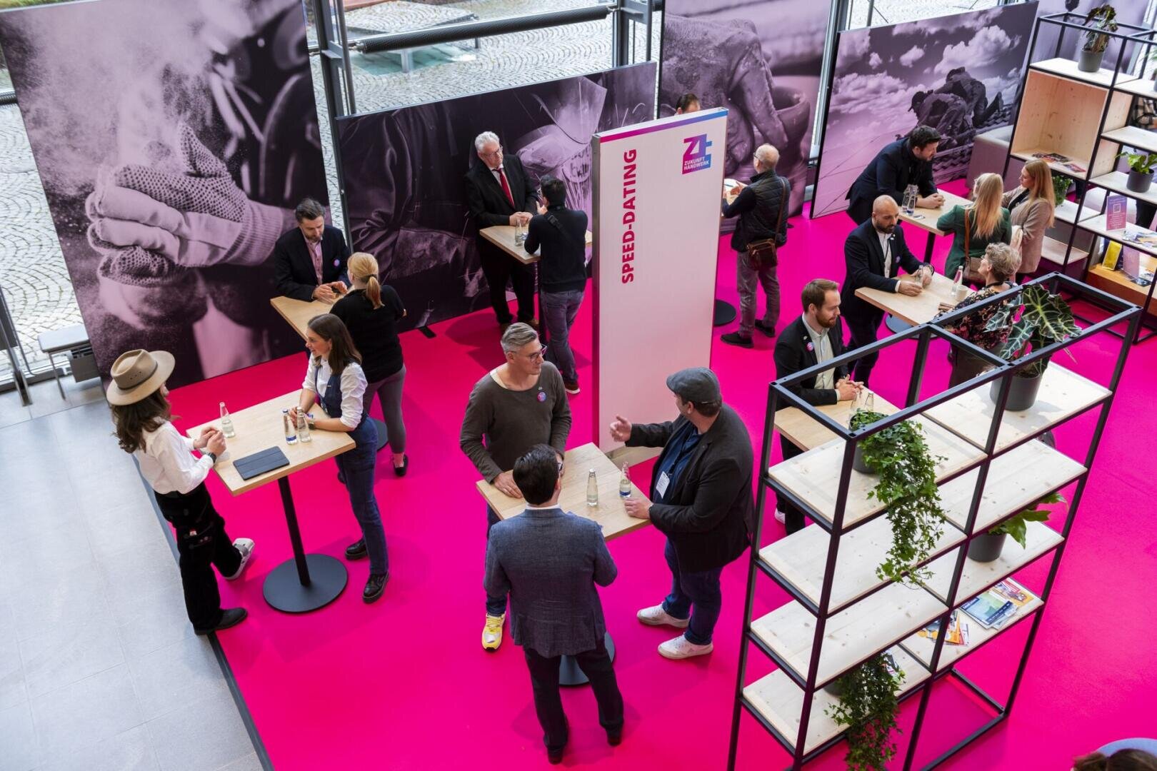 Menschen nehmen an einer Networking- oder Speed-Dating-Veranstaltung teil, stehen paarweise an hohen Tischen auf einem leuchtend rosa Boden und unterhalten sich. Schwarzweißfotos und Pflanzen schmücken den modernen Veranstaltungsort.