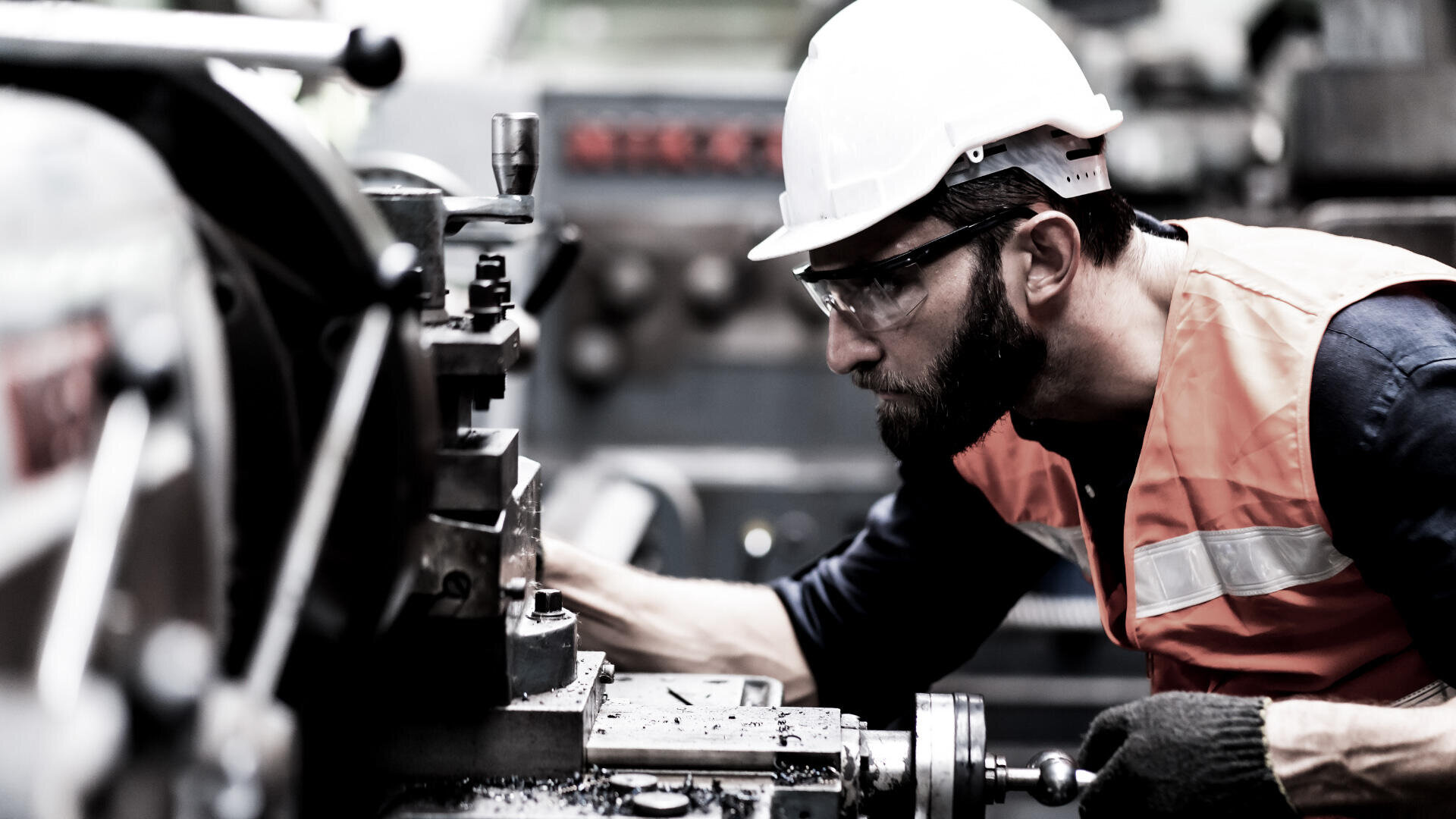 Ein Mann mit weißem Schutzhelm, Schutzbrille und orangefarbener Warnweste bedient Industriemaschinen in einer Fabrik und konzentriert sich voll und ganz auf seine Arbeit.