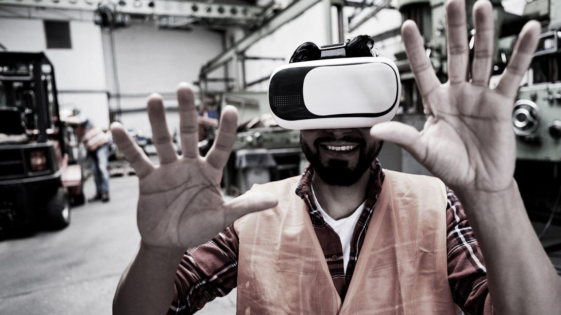 Ein Mann mit Sicherheitsweste und Virtual-Reality-Headset steht lächelnd in einer Industrieumgebung und hat die Hände vor sich erhoben, als würde er mit einer virtuellen Umgebung interagieren.
