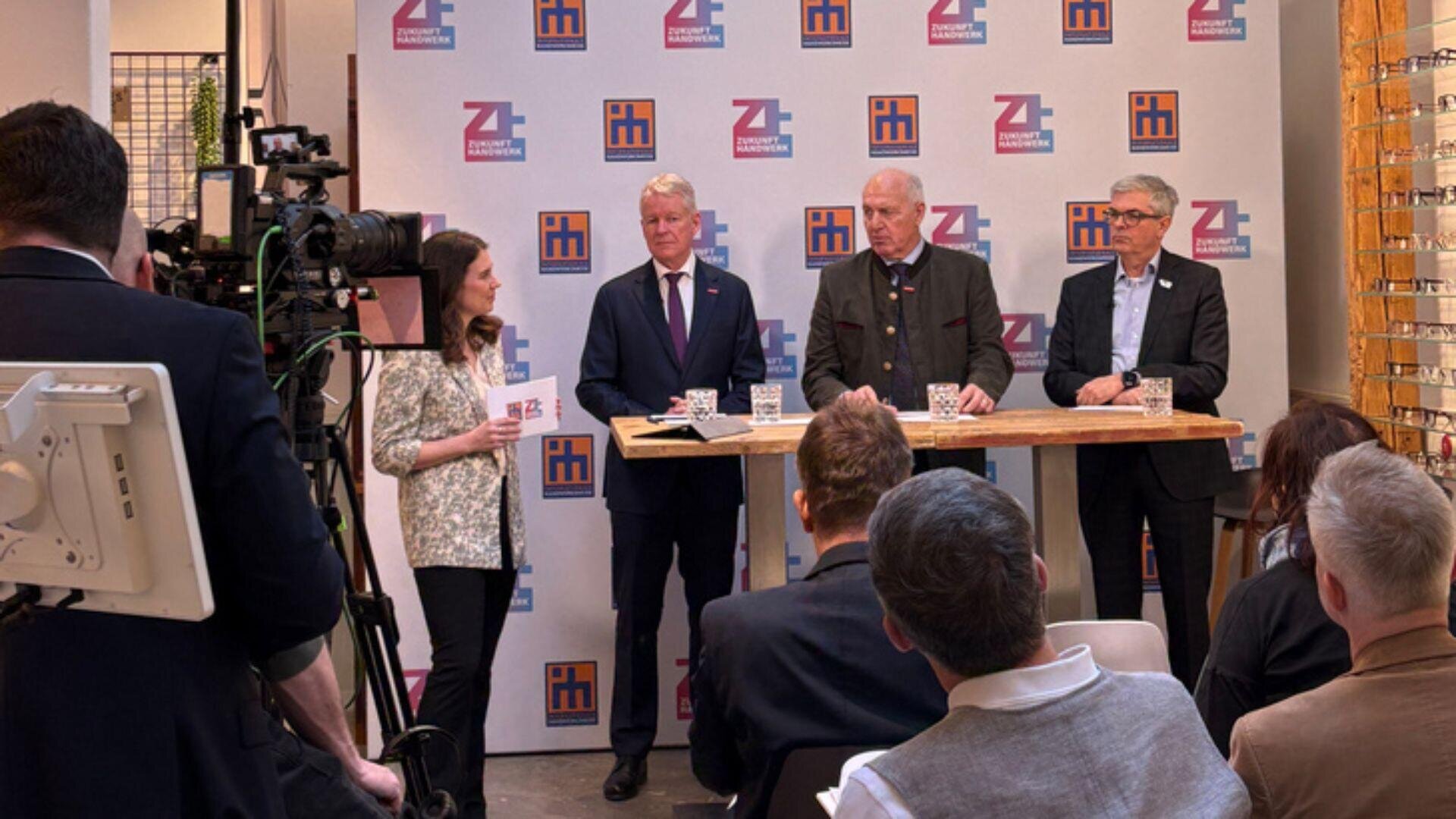 Eine Frau moderiert eine Podiumsdiskussion mit drei Männern in Anzügen, die an einem Stehtisch stehen, während Publikum und Kameras zuschauen. Hinter ihnen befindet sich ein Markenhintergrund mit Logos.