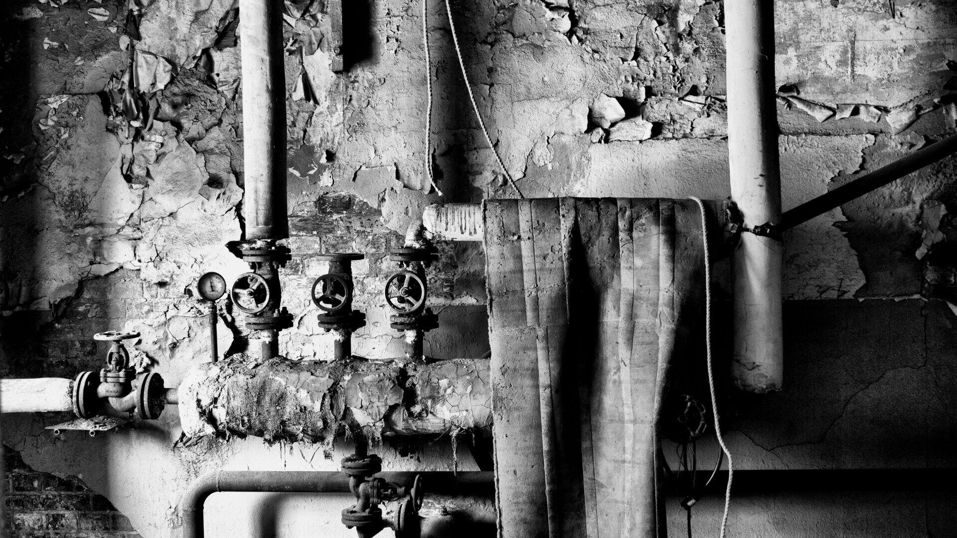 Schwarz-Weiß-Foto alter, verrosteter Rohre und Ventile vor einer abblätternden, rissigen Wand. Ein alter, gefalteter Stoffschlauch hängt über den Rohren und suggeriert Verlassenheit und Verfall.