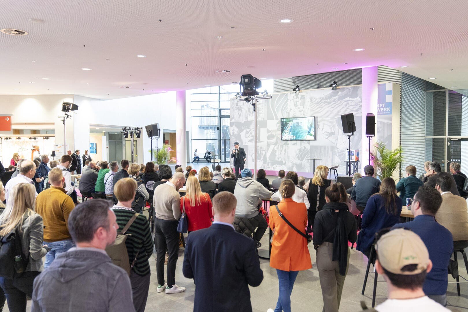 Eine Menschenmenge sitzt und steht einem Redner auf einer Bühne in einem modernen Innenraum gegenüber. Hinter dem Redner befinden sich eine Leinwand und Lichter, und große Fenster lassen natürliches Licht herein.