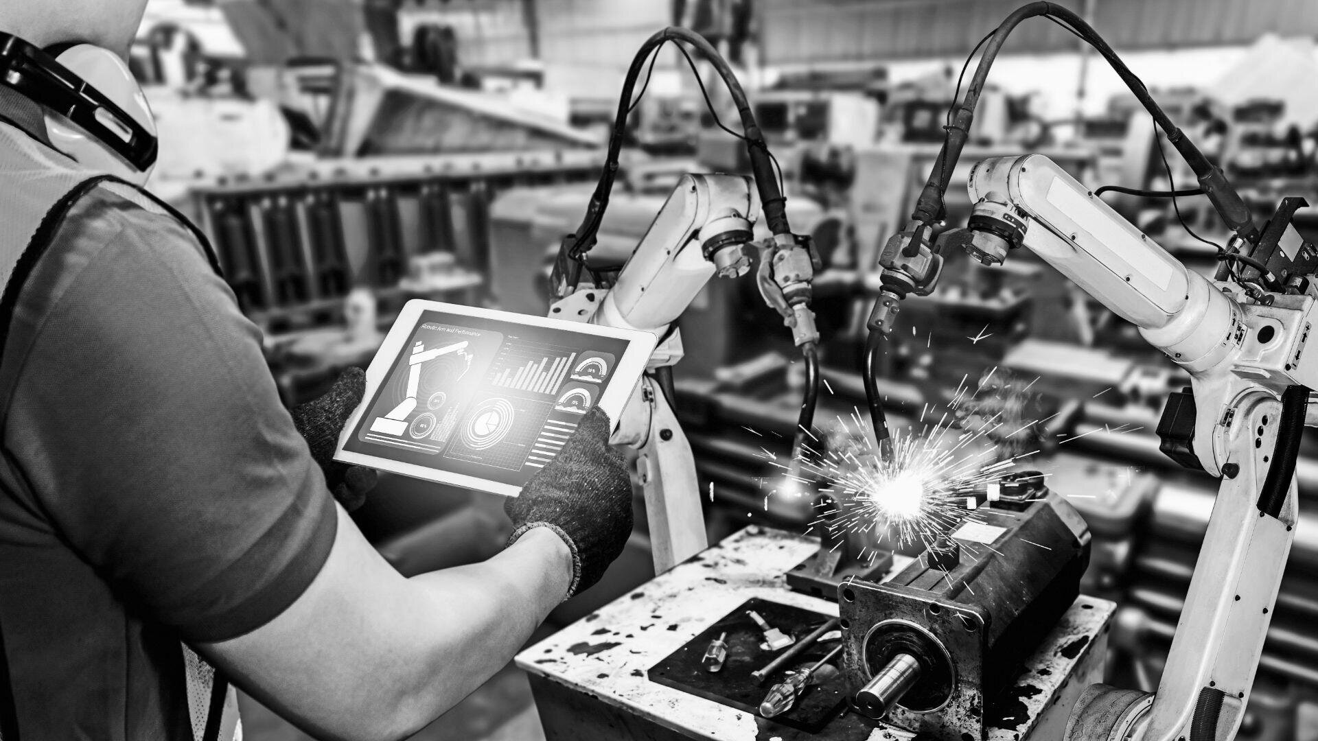 Eine Person mit Handschuhen hält ein Tablet mit Datenanzeige und überwacht gleichzeitig Roboterarme, die in einer Fabrik Schweißarbeiten durchführen.