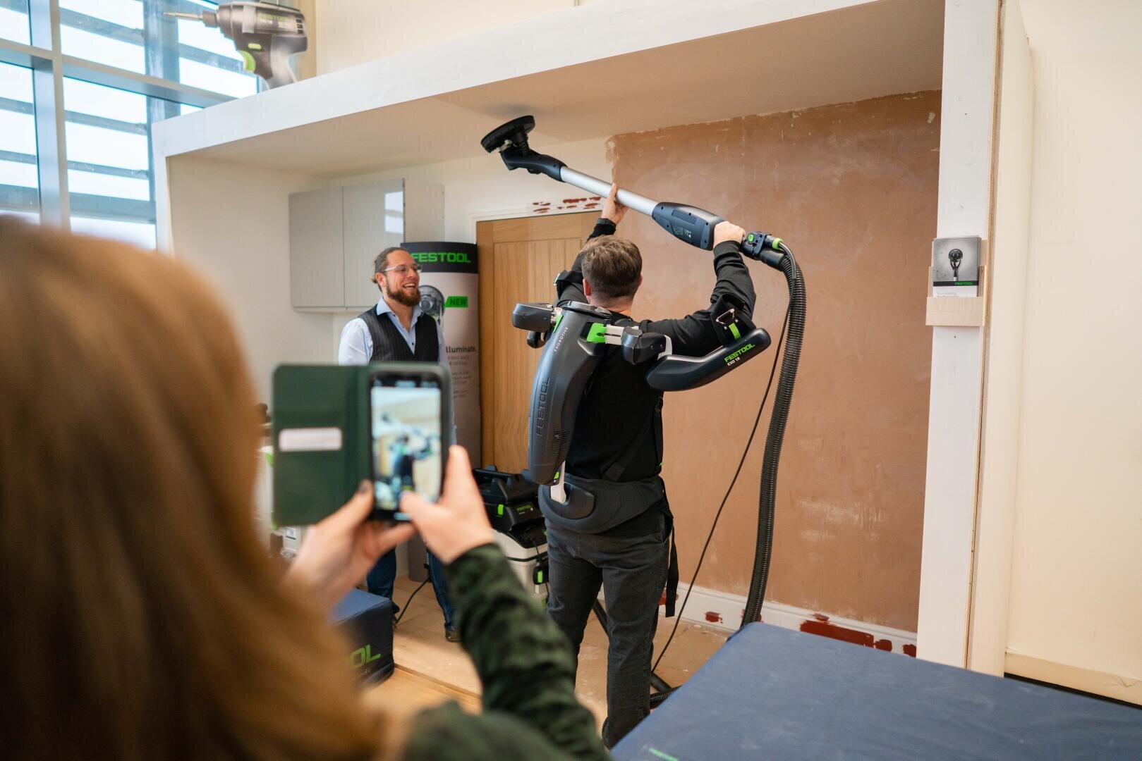 Eine Person, die ein Exoskelett trägt, saugt eine hohe Wand, während eine andere Person ein Foto mit einem Smartphone macht; eine dritte Person schaut in einem teilweise unfertigen Raum zu.