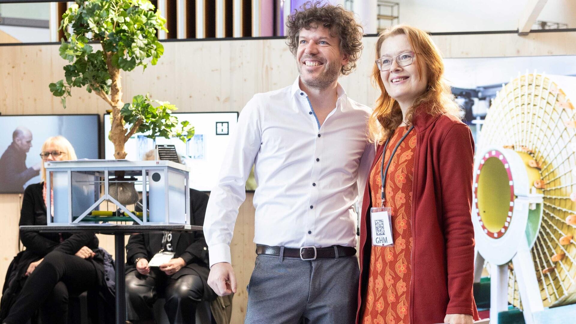 Ein lächelnder Mann und eine lächelnde Frau stehen bei einer Indoor-Veranstaltung zusammen und posieren für ein Foto. Sie stehen vor einer blauen Maschine und Ausstellungsdisplays. Die Frau hält eine Tragetasche in der Hand und trägt ein Veranstaltungsabzeichen um den Hals.